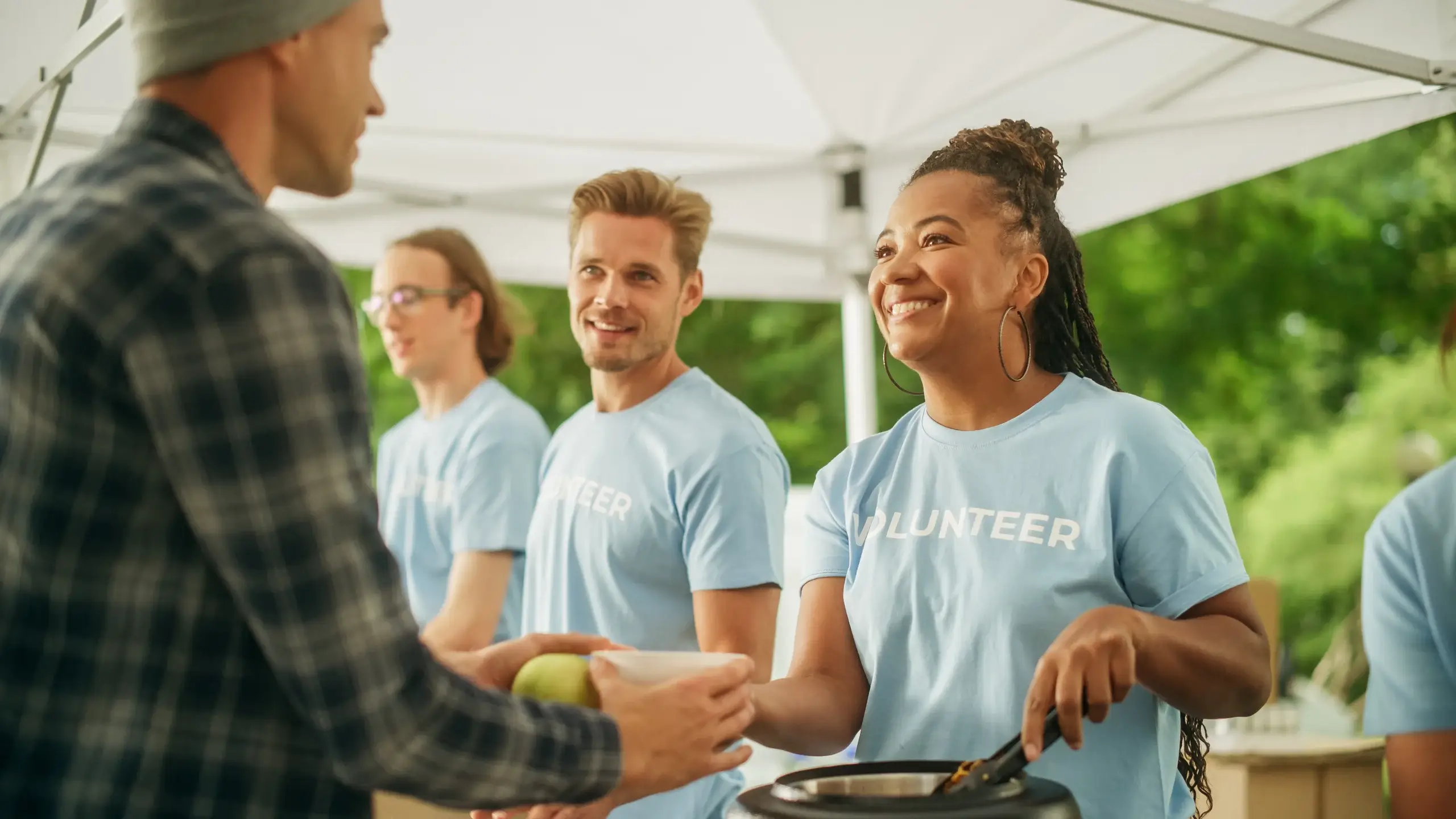 Volunteers in light blue shirts serve food at an outdoor event. A woman with braided hair smiles while handing a bowl to a man. Two other volunteers stand nearby under a white canopy.