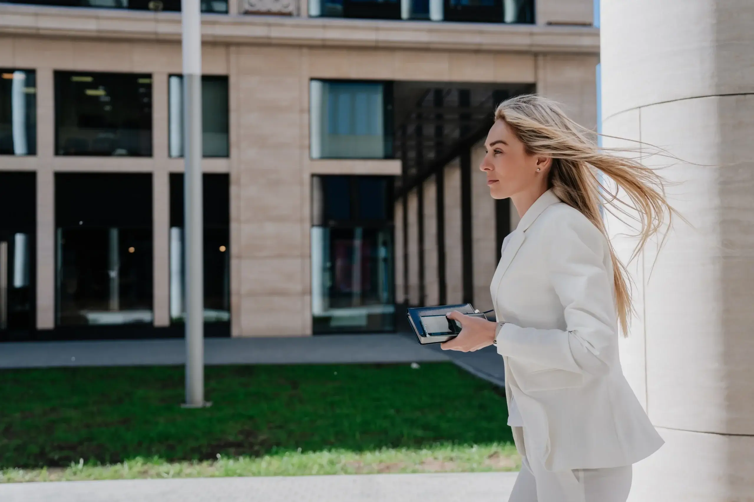 A woman with long blonde hair walks outdoors by a modern building. She is holding a tablet and dressed in a white suit. The background shows a multi-story structure with large windows and a patch of grass. Her hair flows in the wind.