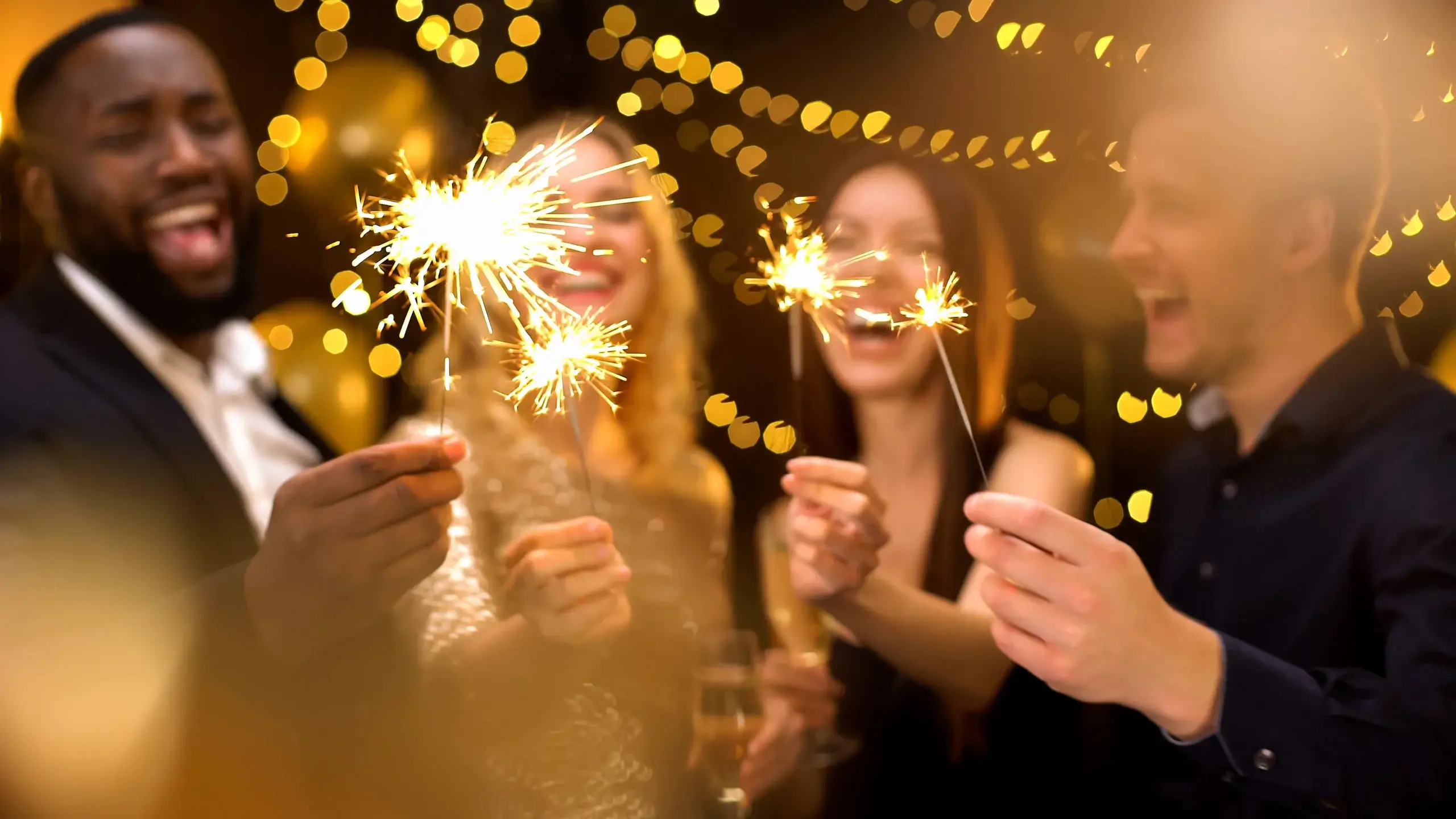 Four people smiling and holding sparklers, surrounded by warm, blurred string lights in the background. The scene suggests a festive celebration, with everyone appearing joyful and engaged in the moment.