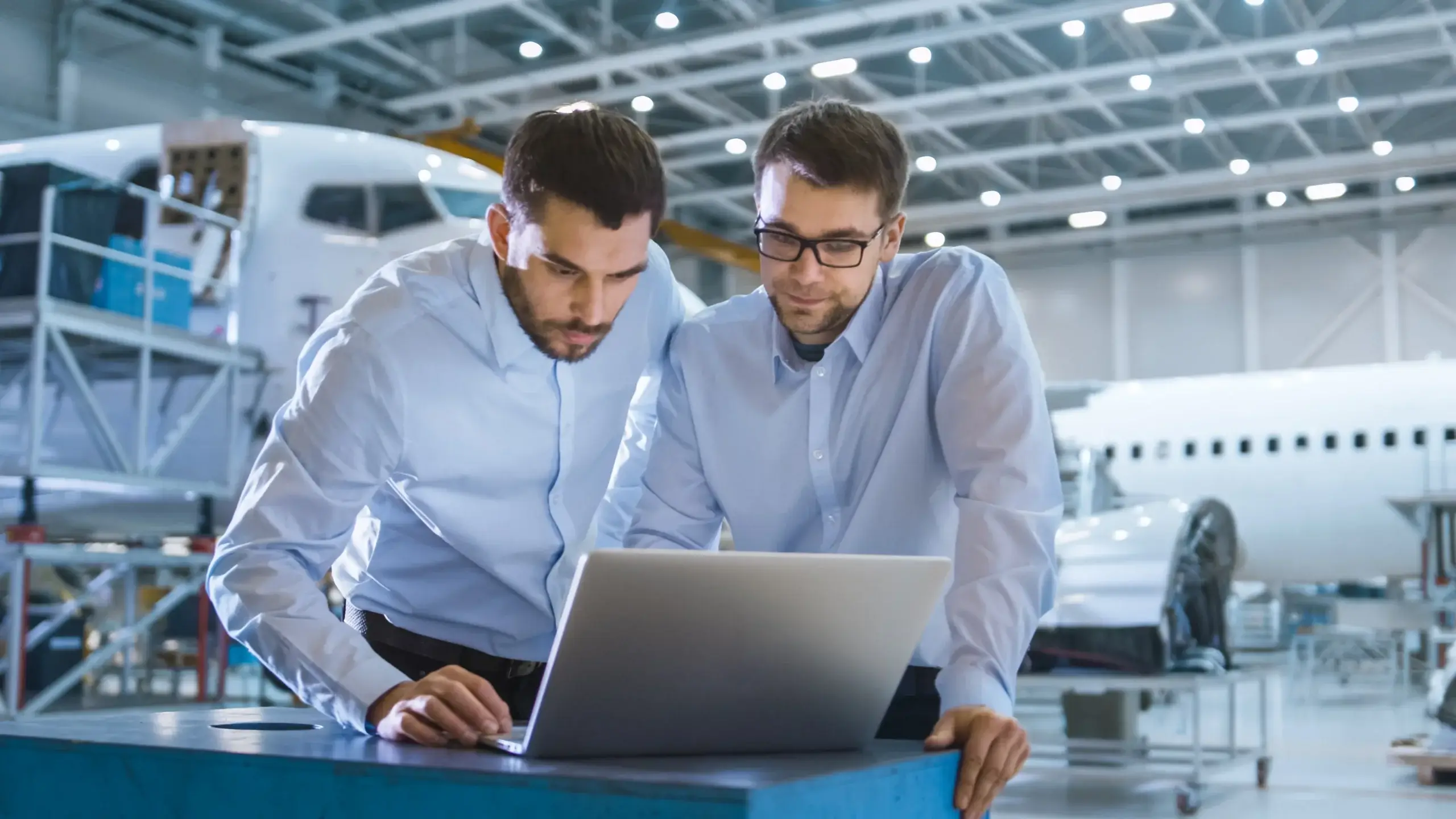 Two men in blue shirts look at a laptop inside an aircraft hangar. They seem focused and are surrounded by airplane parts and equipment, suggesting an engineering or maintenance context.