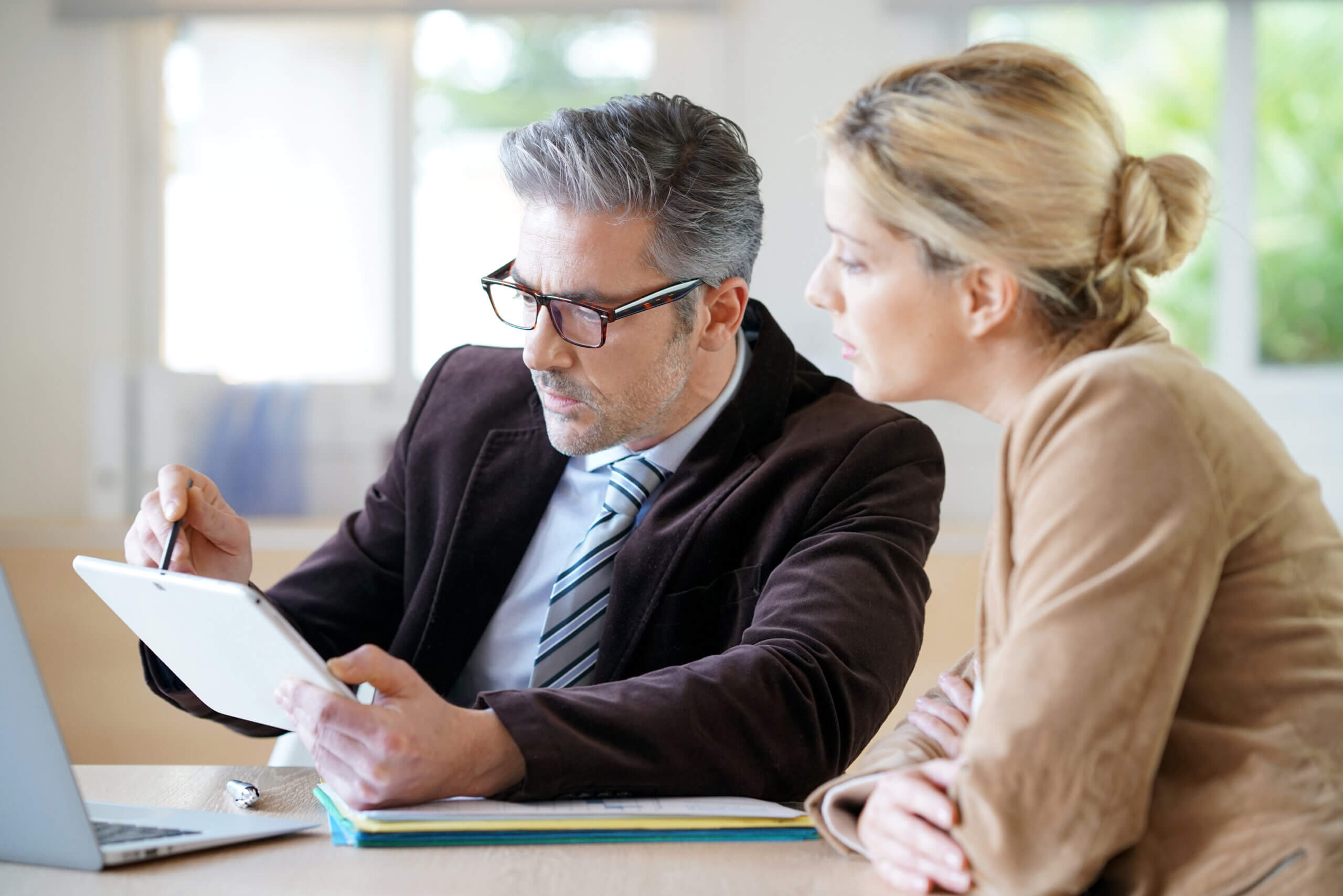 A man with glasses and gray hair is showing a tablet to a woman with blonde hair. They are both seated at a table with a laptop and documents in a well-lit room. The woman is attentively looking at the screen.