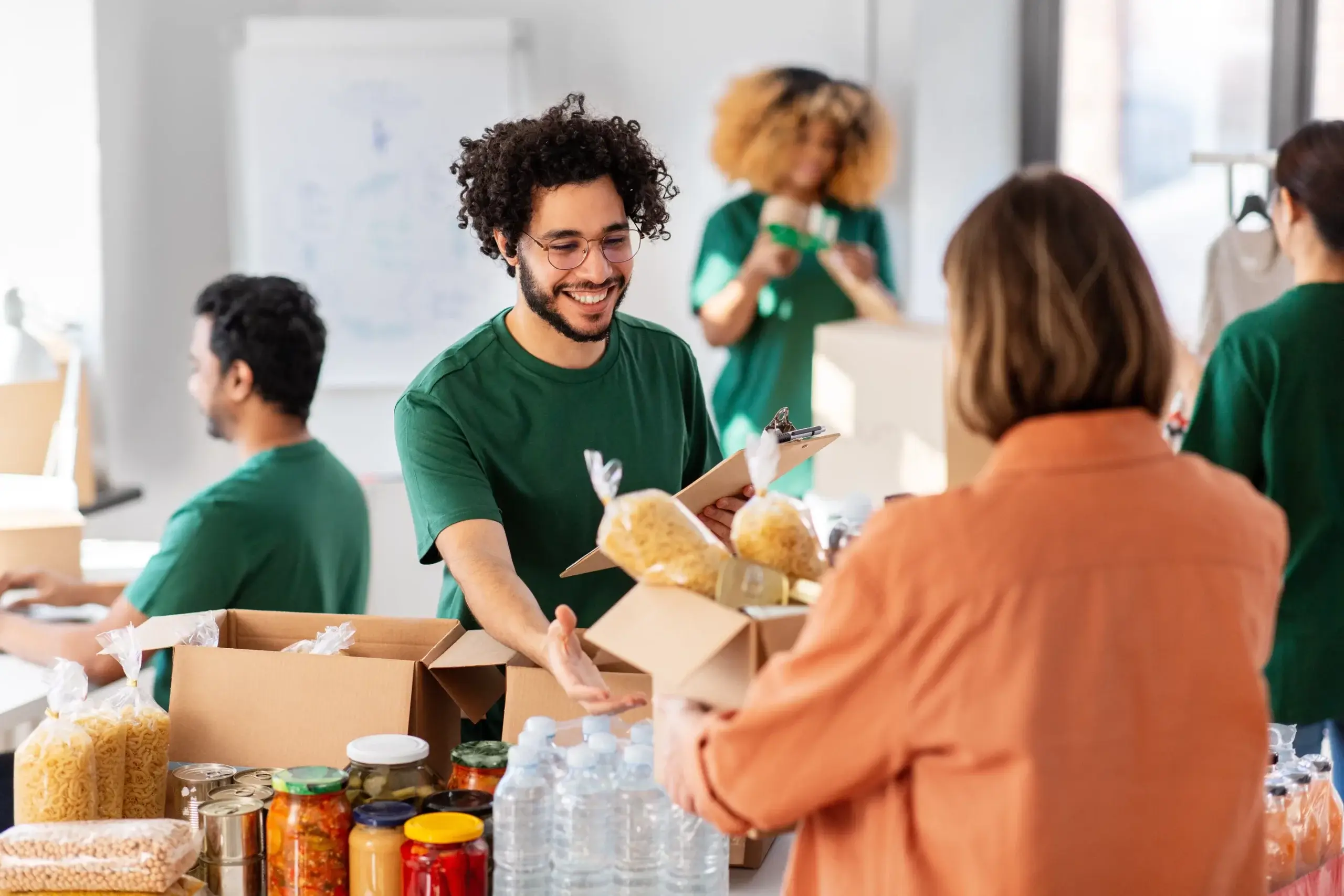 A man with curly hair and glasses hands a box containing pasta to a woman in an orange shirt. They are part of a group packing food items, including jars and water bottles, in a bright room. Another person works in the background.