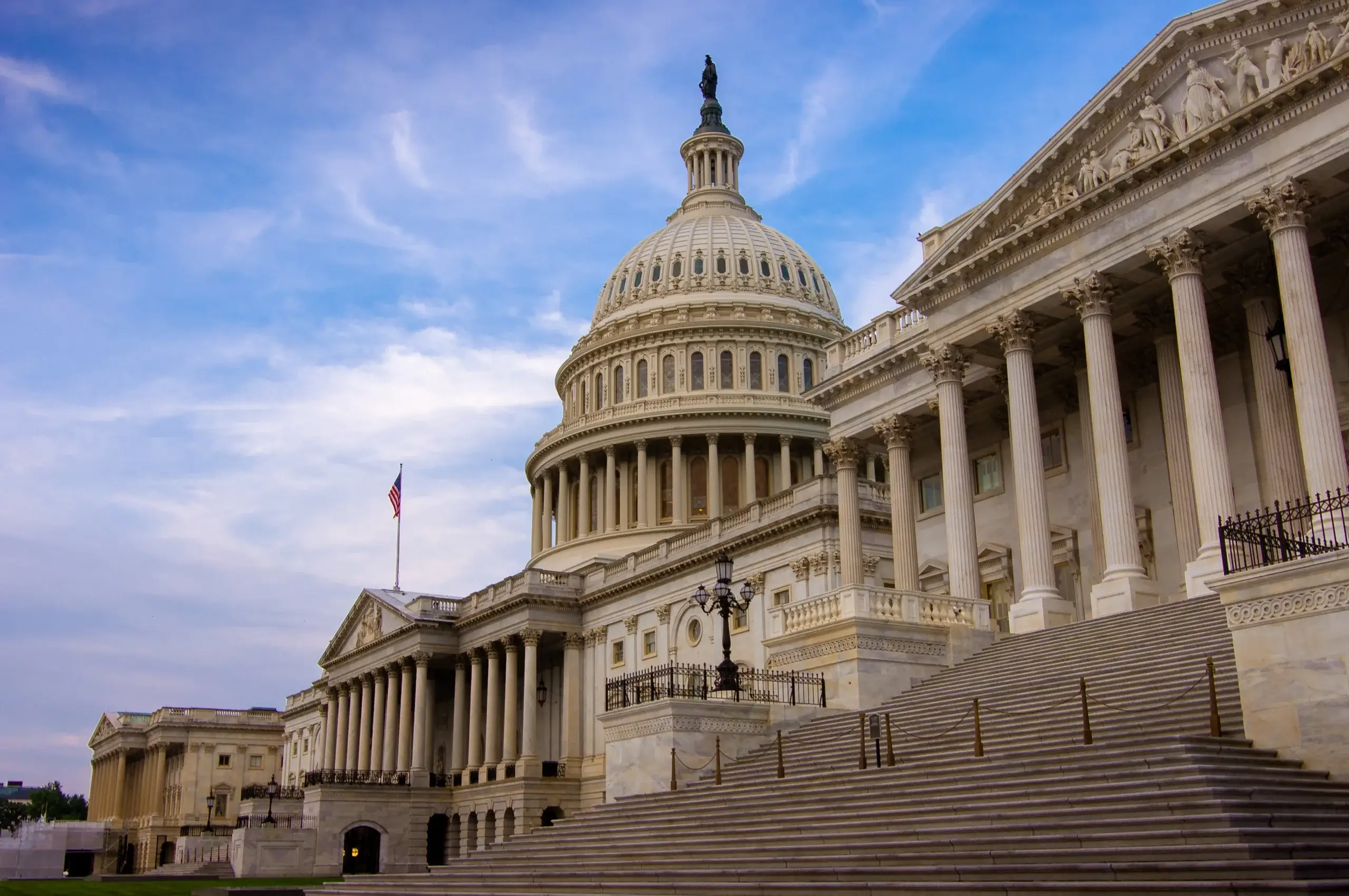 The image shows the United States Capitol building in Washington, D.C., featuring its iconic dome and columns. A cloudy sky forms the backdrop, and an American flag is visible near the structure.