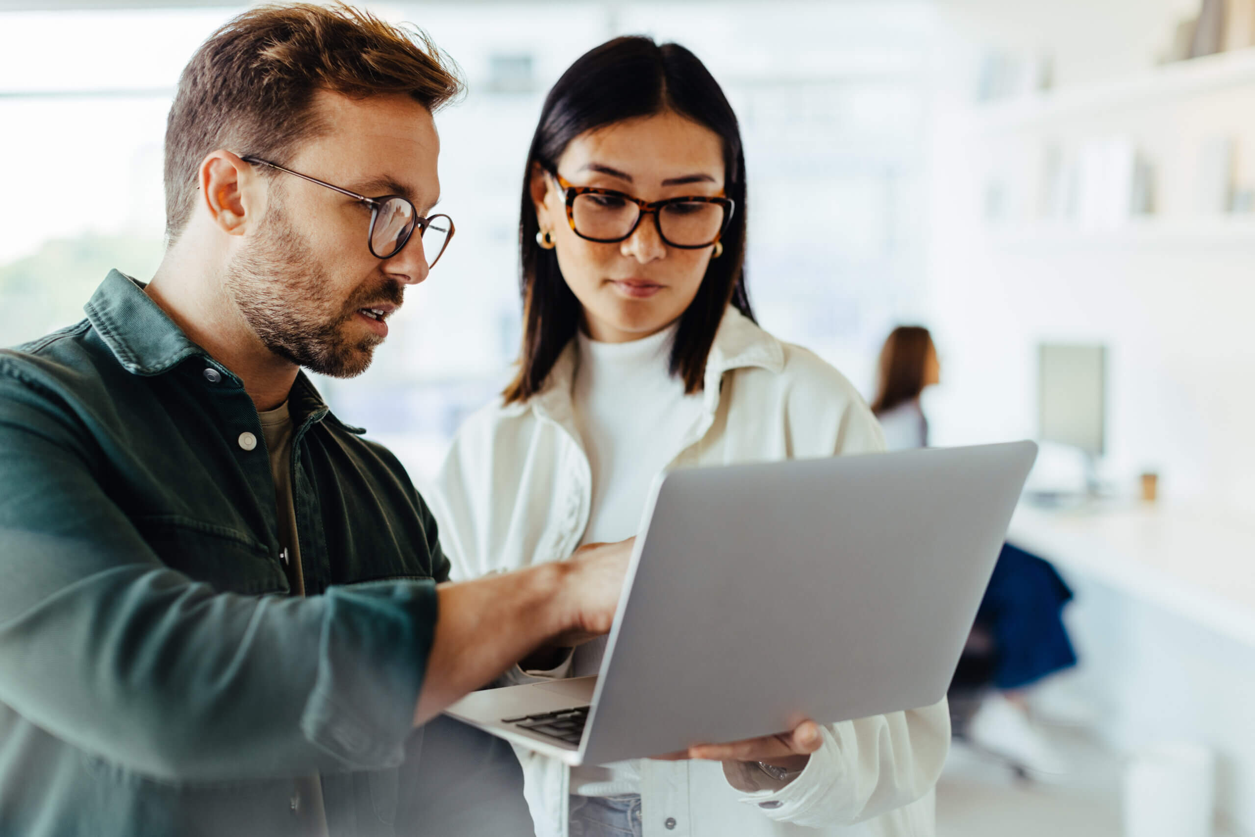 A man and woman in glasses are standing and looking at a laptop screen. The man is pointing at the screen while the woman holds the laptop. They are in a bright office space with shelves and another person in the background.