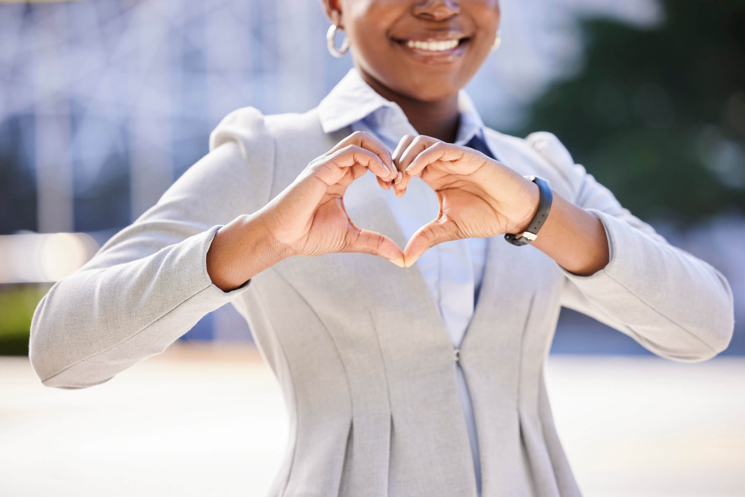 A person in a light gray blazer makes a heart shape with their hands. They are smiling softly, standing outdoors with a blurred background.