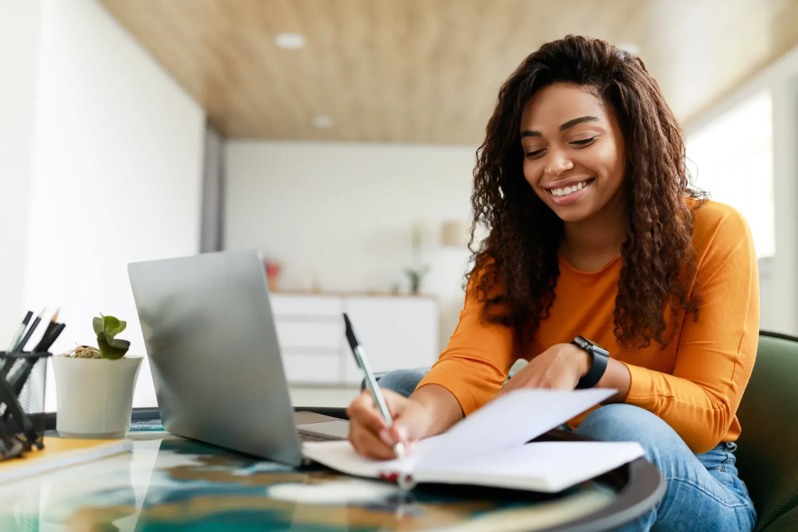 A woman with curly hair, wearing an orange sweater, sits at a table writing in a notebook. She's smiling and looking at a laptop. The room is bright, with a plant and a blurred background.
