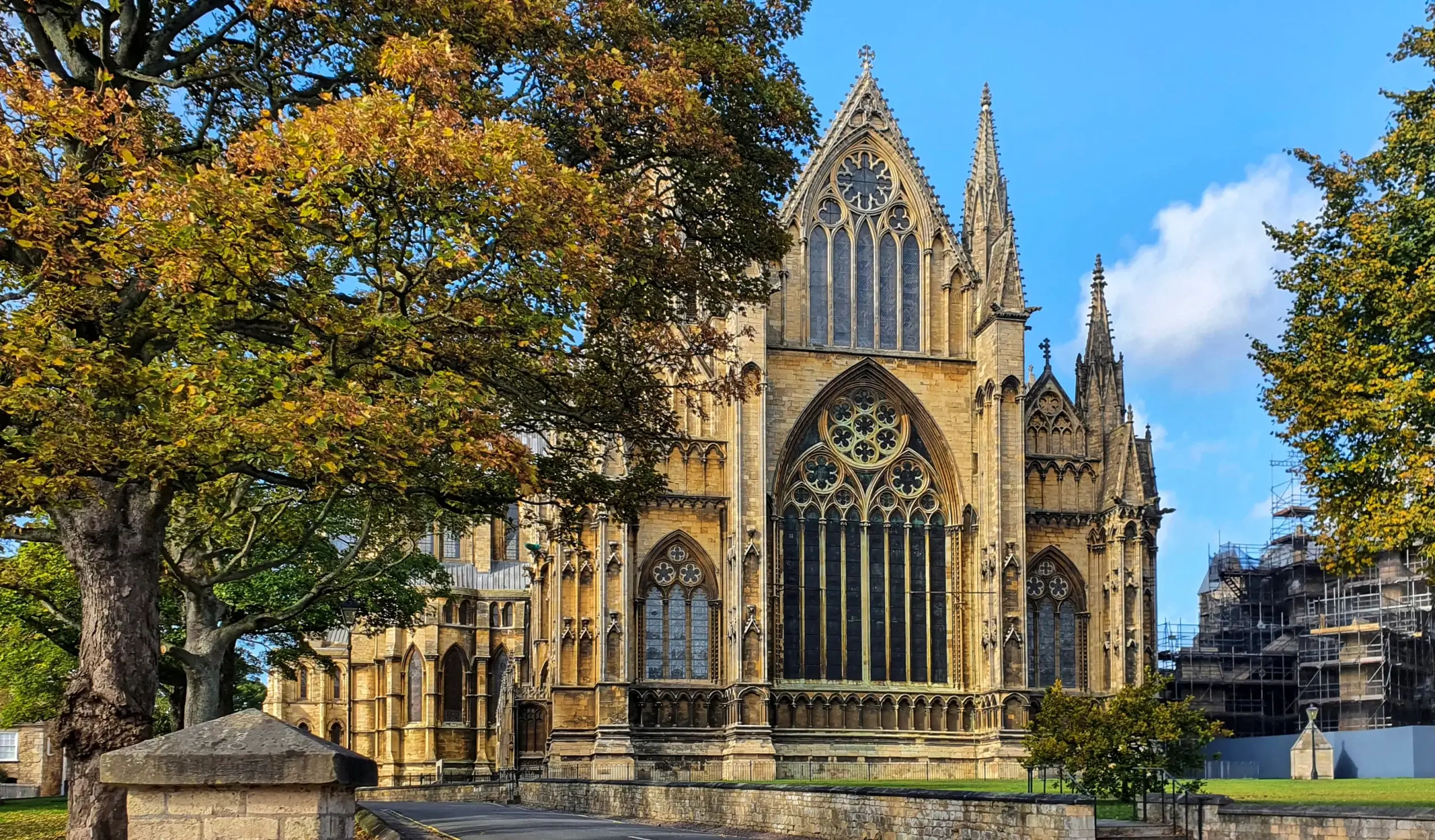 A historic Gothic cathedral with intricate details and large stained glass windows, surrounded by autumn trees. The sky is clear and blue, creating a picturesque scene. Scaffolding is visible on the right side of the building.