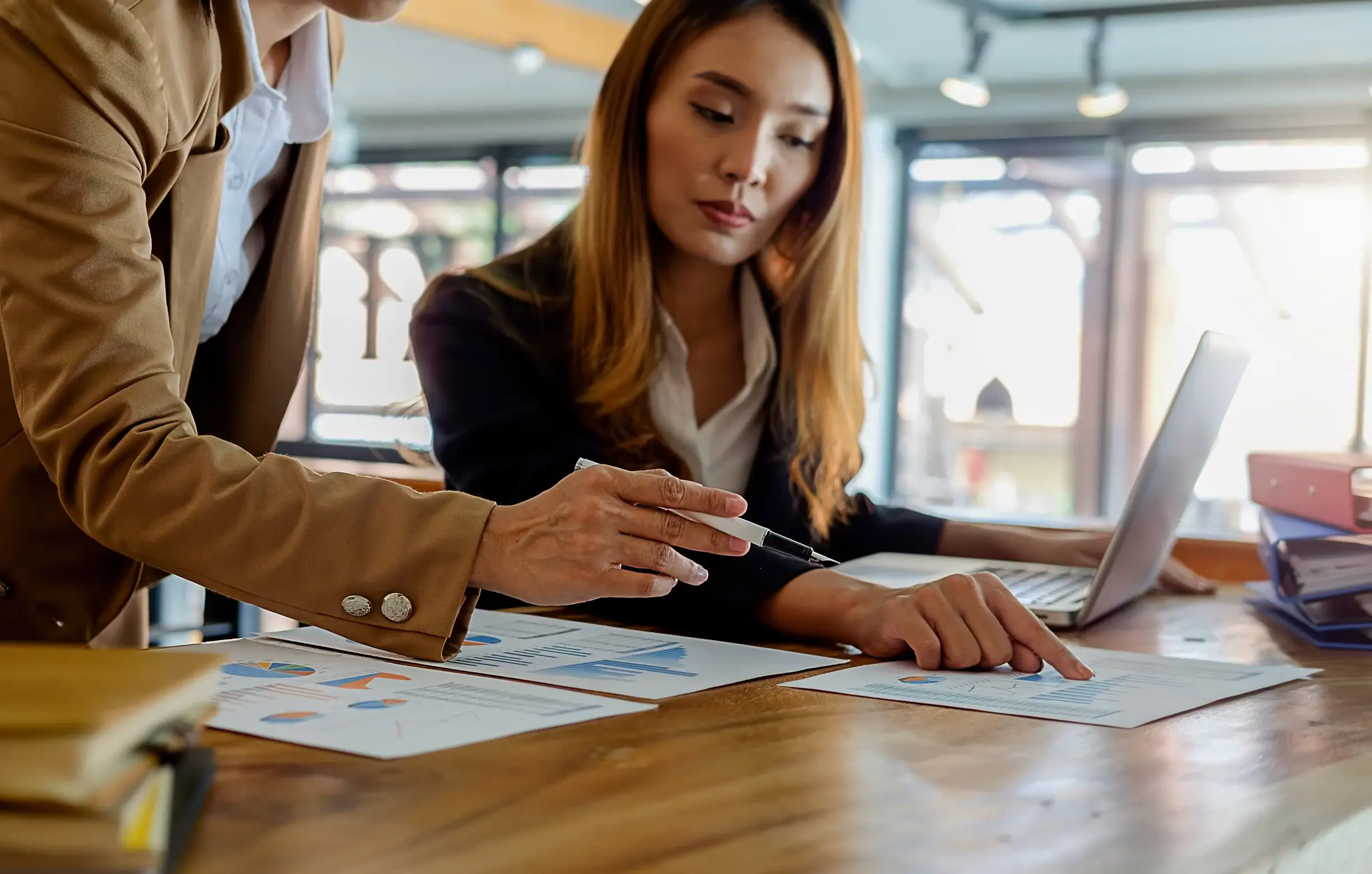 Two professionals in an office setting review documents with charts and graphs on a wooden table. One points with a pen, while the other looks attentively. A laptop and stack of binders are in the background.