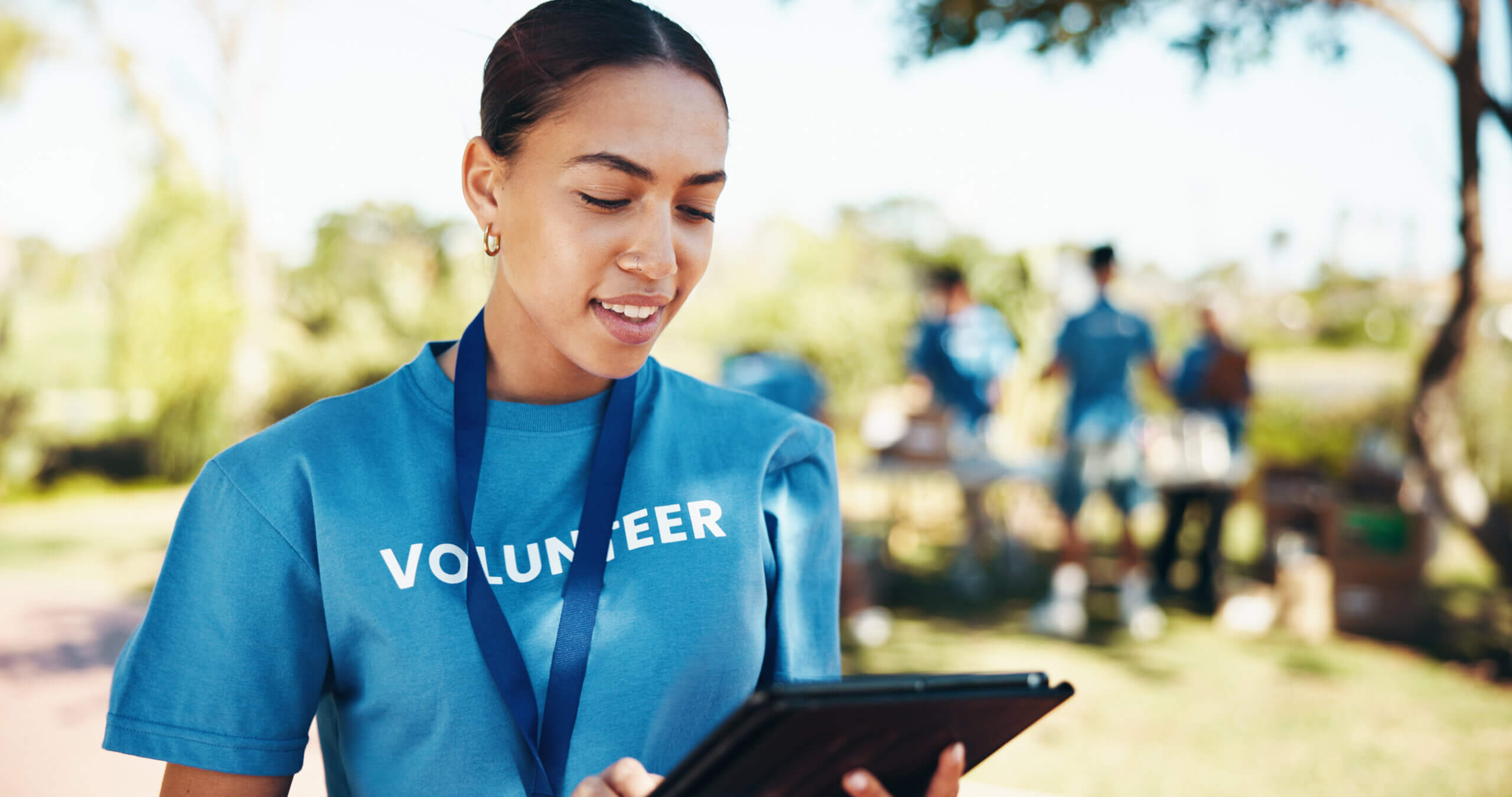 A woman wearing a blue "Volunteer" shirt and a lanyard holds a tablet outside. The background shows other people in blue shirts involved in an outdoor activity, with trees and bright sunlight.