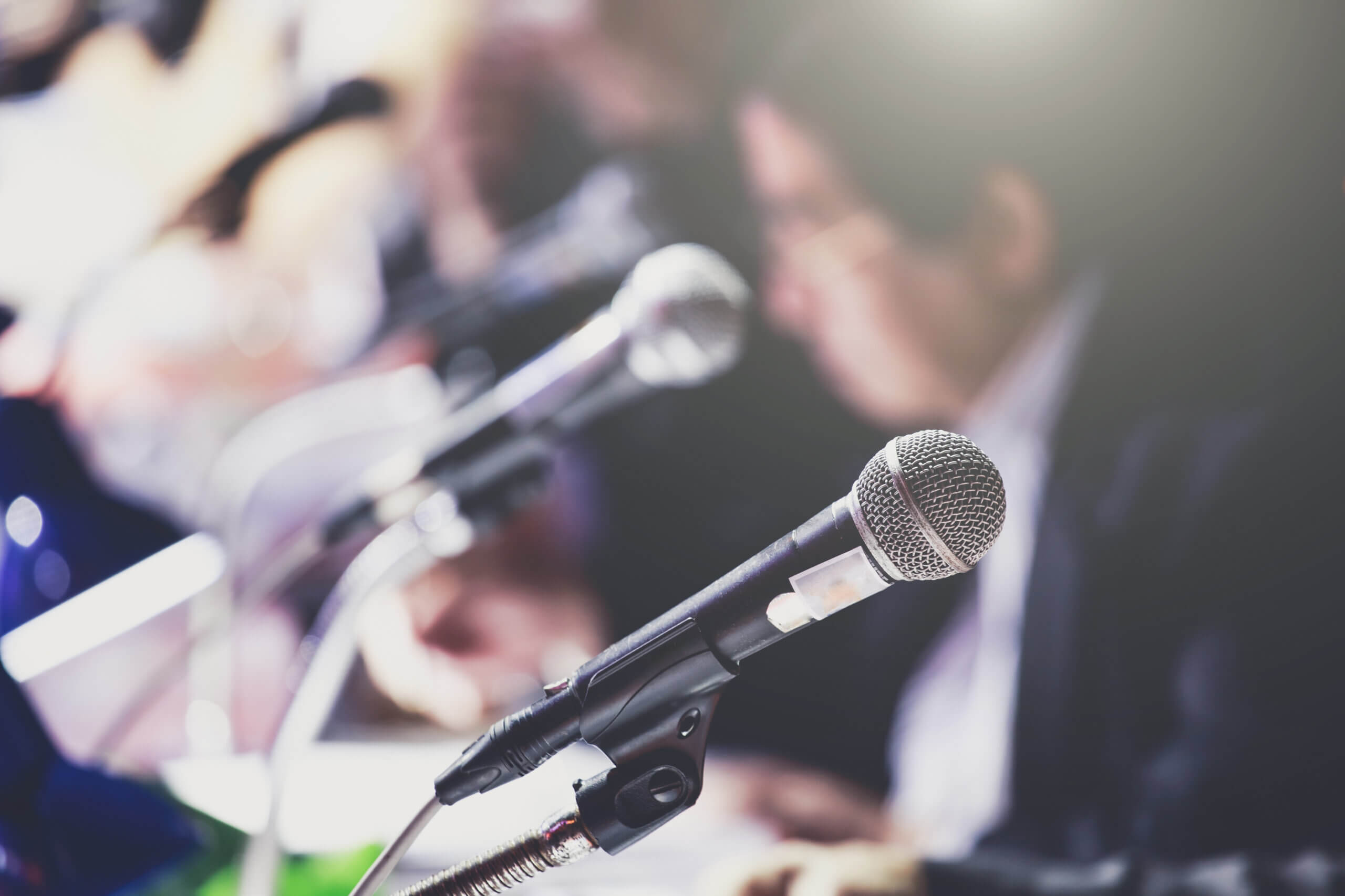Close-up of a microphone at a conference table. Several other microphones sit in a row, with papers and blurred people in the background. Soft lighting suggests a formal setting or press event.