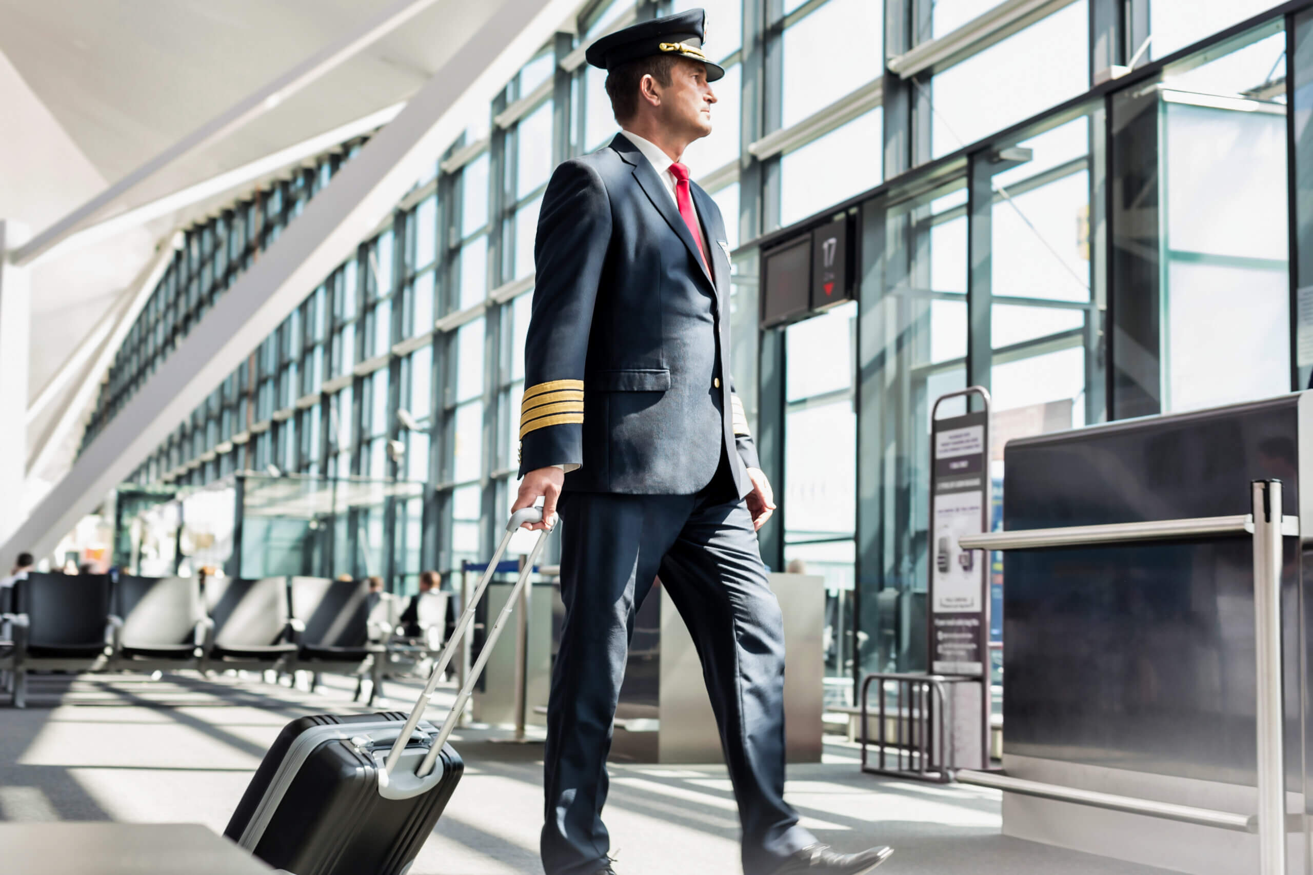 Airline pilot in uniform, walking through an airport terminal, pulling a rolling suitcase. The terminal has large windows allowing natural light. The pilot is wearing a cap, a dark suit with gold stripes on the sleeves, and a red tie.