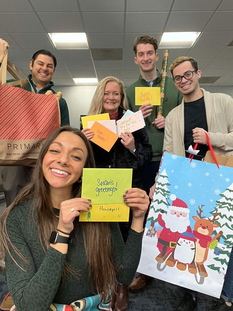 A group of four people from Cogency Global, smiling and holding holiday cards and gift bags. One gift bag has a Santa Claus and penguin design. Casually dressed, they stand in a room with a checkered ceiling and fluorescent lights, embodying the spirit of compliance services camaraderie.