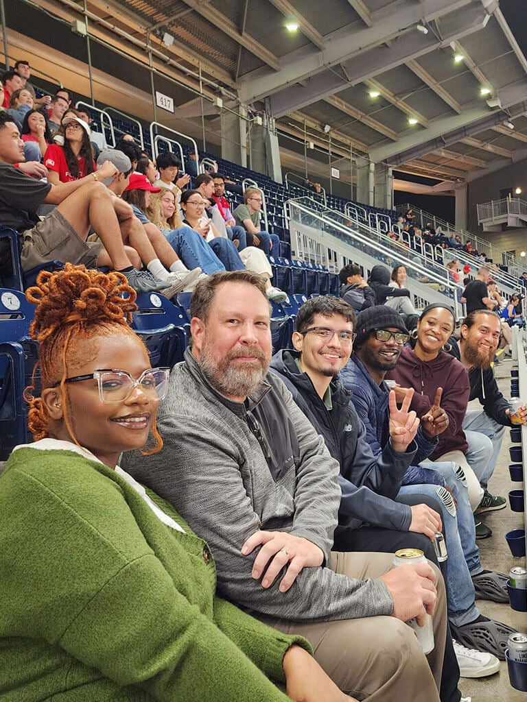 A group of six people, collaborating on corporate compliance services at a sports stadium, sit together smiling. Some hold drinks, and the background reveals partially filled stands with other attendees. The atmosphere is cheerful and relaxed, reminiscent of a seamless partnership with Cogency Global.