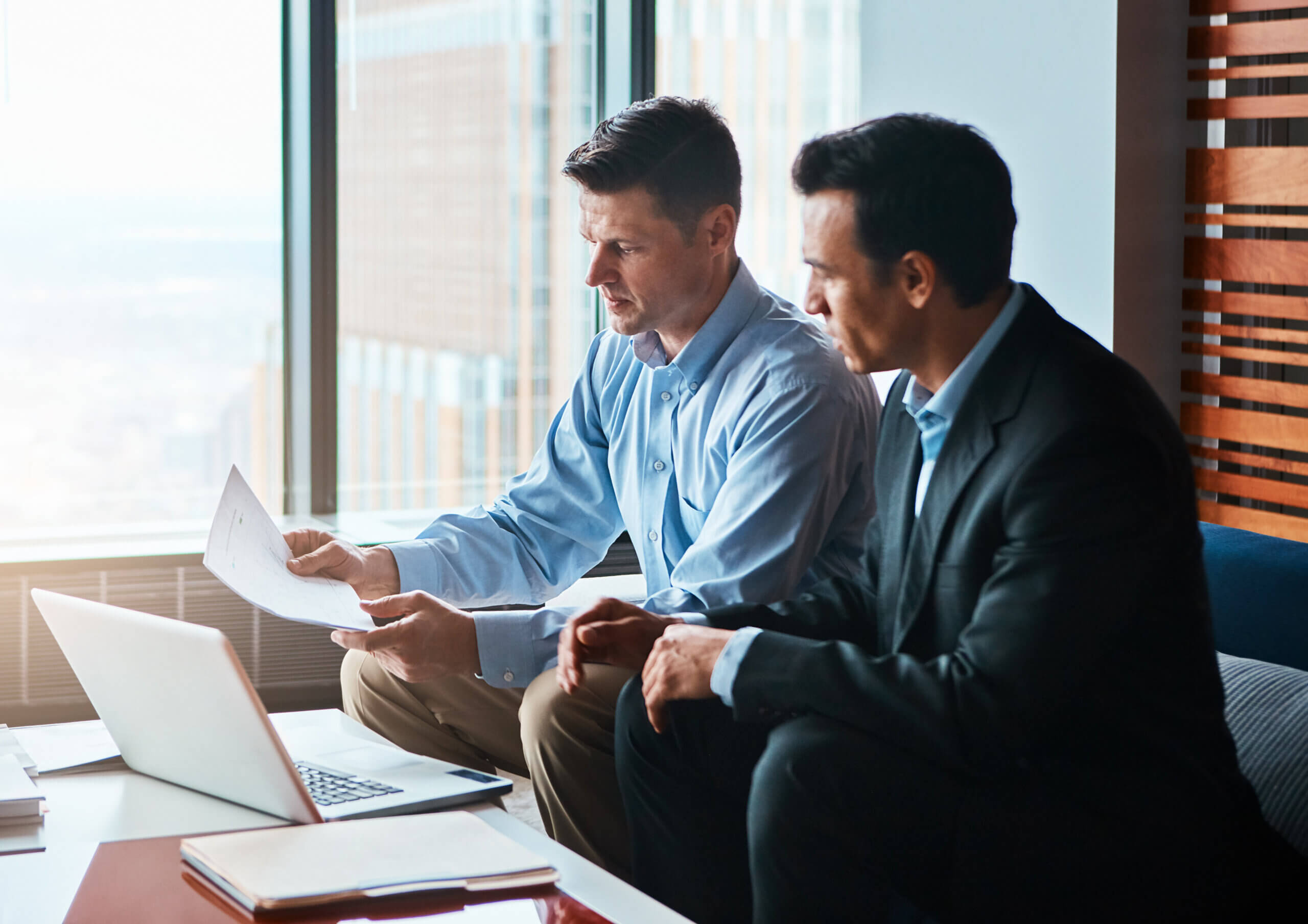 Two men in business attire sit in a modern office. One holds documents, and both focus intently on them. A laptop is open on the table, with large windows offering a city view in the background.