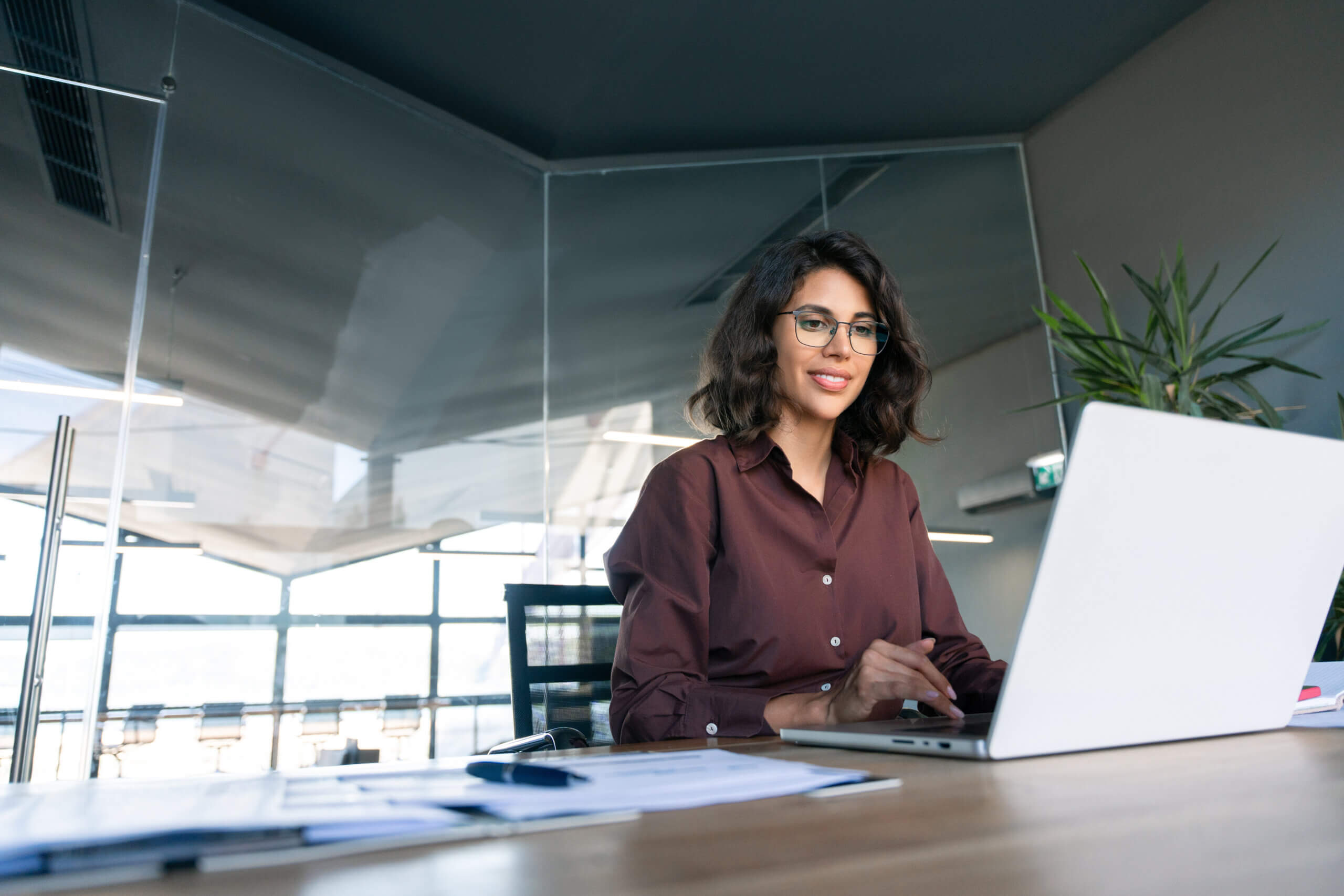 A woman with glasses, wearing a brown blouse, is smiling as she types about Beneficial Ownership Information on her laptop at an office desk. A large window and indoor plant are in the background, enhancing the modern workspace vibe.