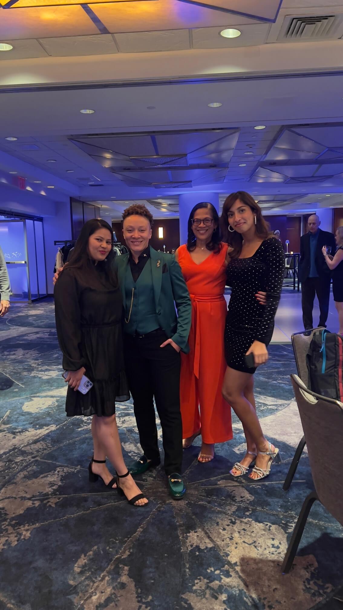 Four people pose for a group photo at what appears to be a formal event. The well-lit, carpeted room hosts three women in black, orange, and black dresses alongside one person in a teal blazer and black pants, perhaps celebrating success with Cogency Global Careers.