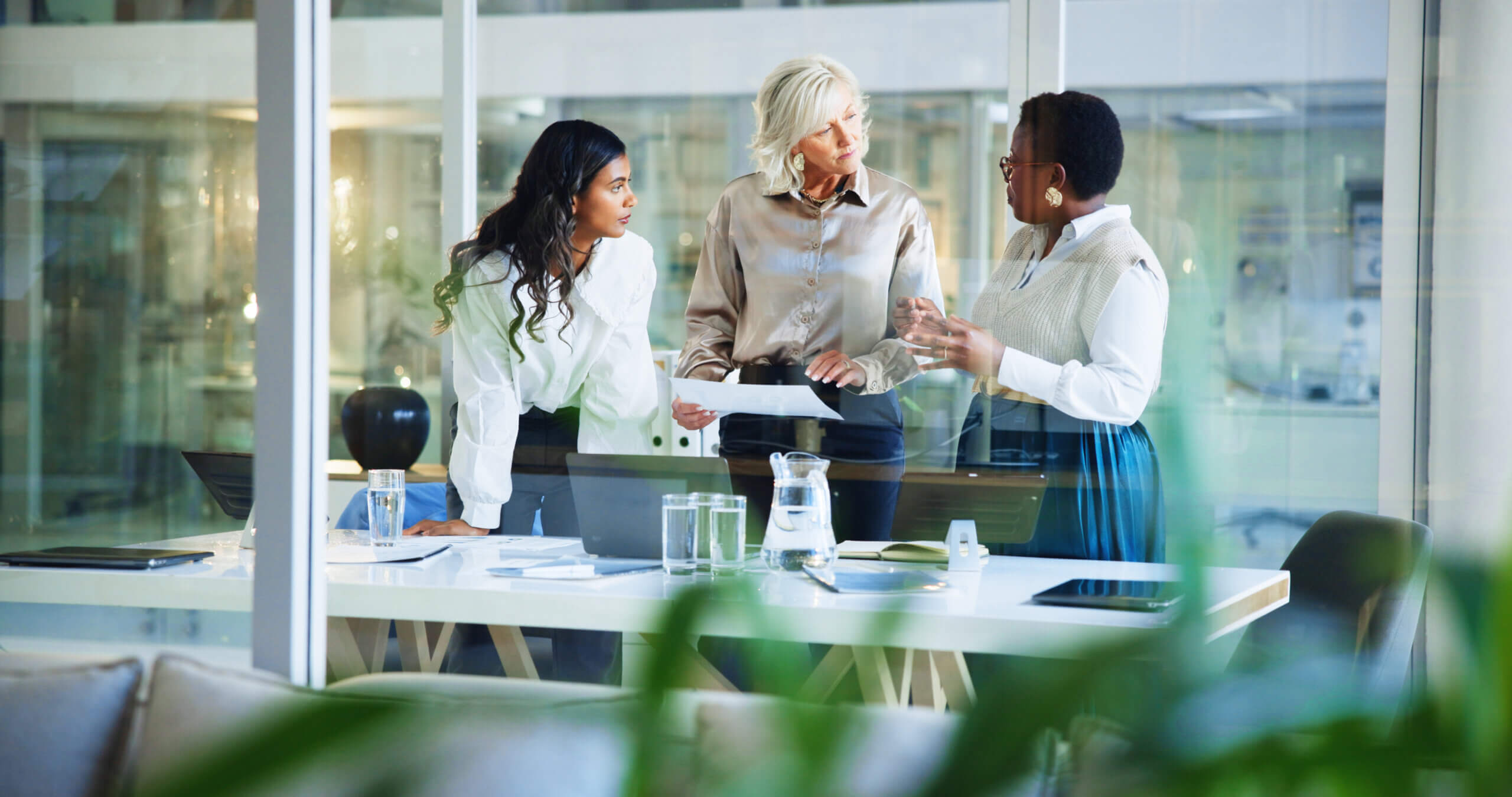 Three professionally dressed women stand and converse around a modern office desk with documents, a laptop, and water glasses, seen through a glass wall with plants in the foreground.