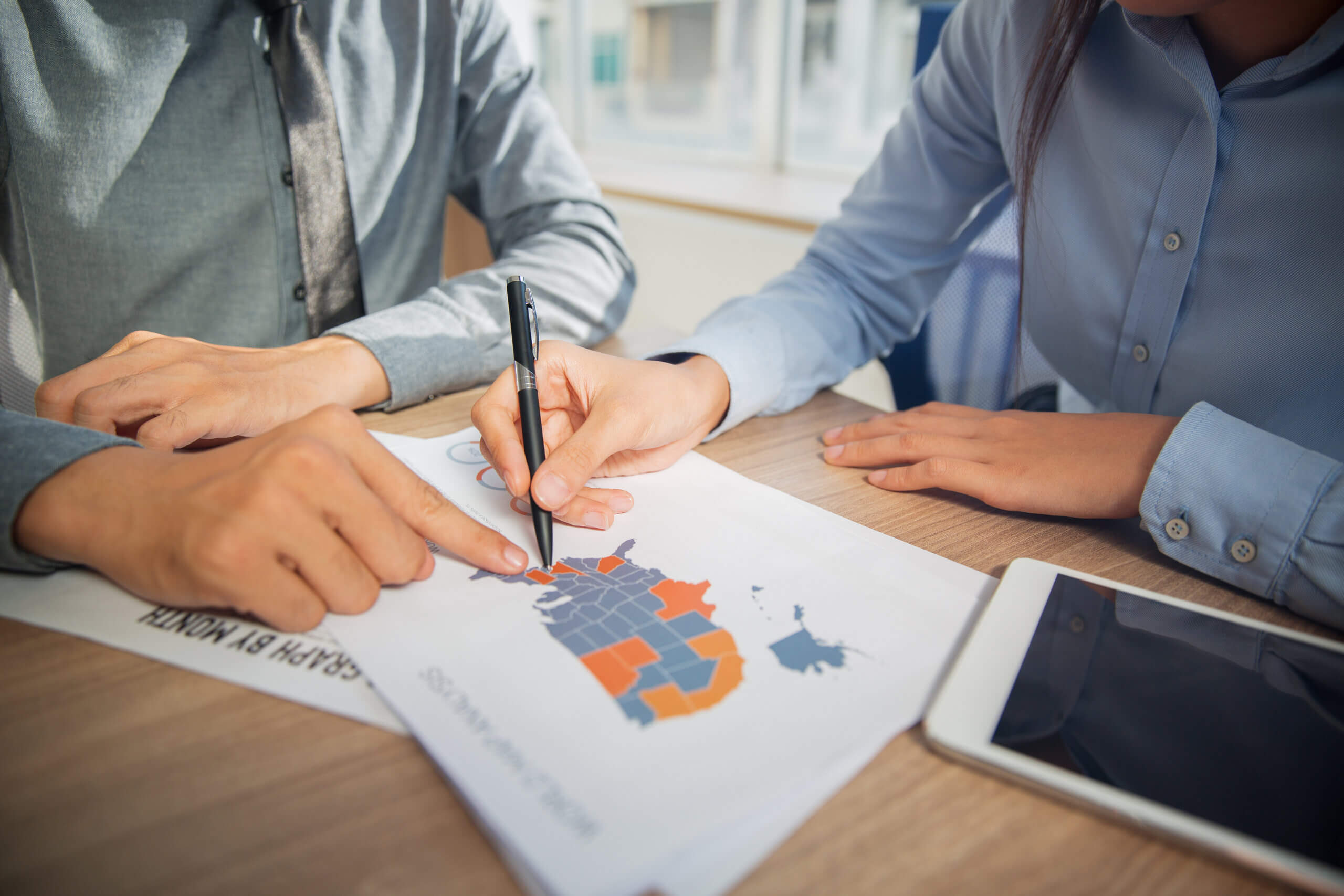 Two people sit at a desk reviewing a document with a colored map of the United States, with one person pointing at the map with a pen. A tablet and other papers are also on the desk.