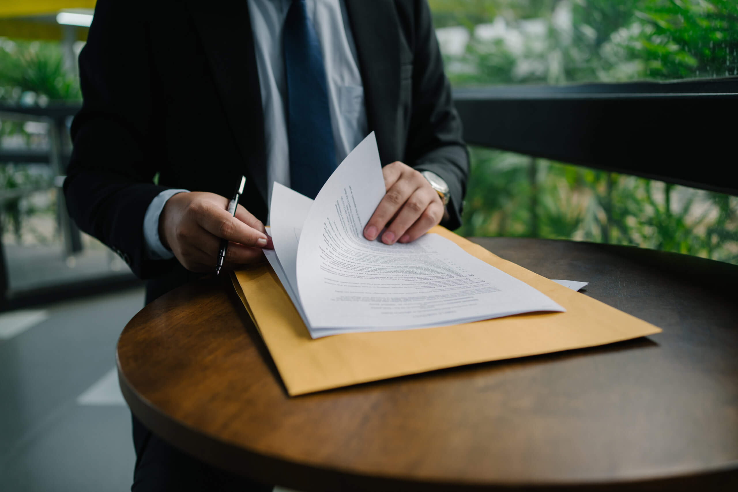 A person in a suit sits at a round table, holding a pen and reviewing documents related to annual report compliance placed on a large manila envelope, with greenery visible outside the window in the background.
