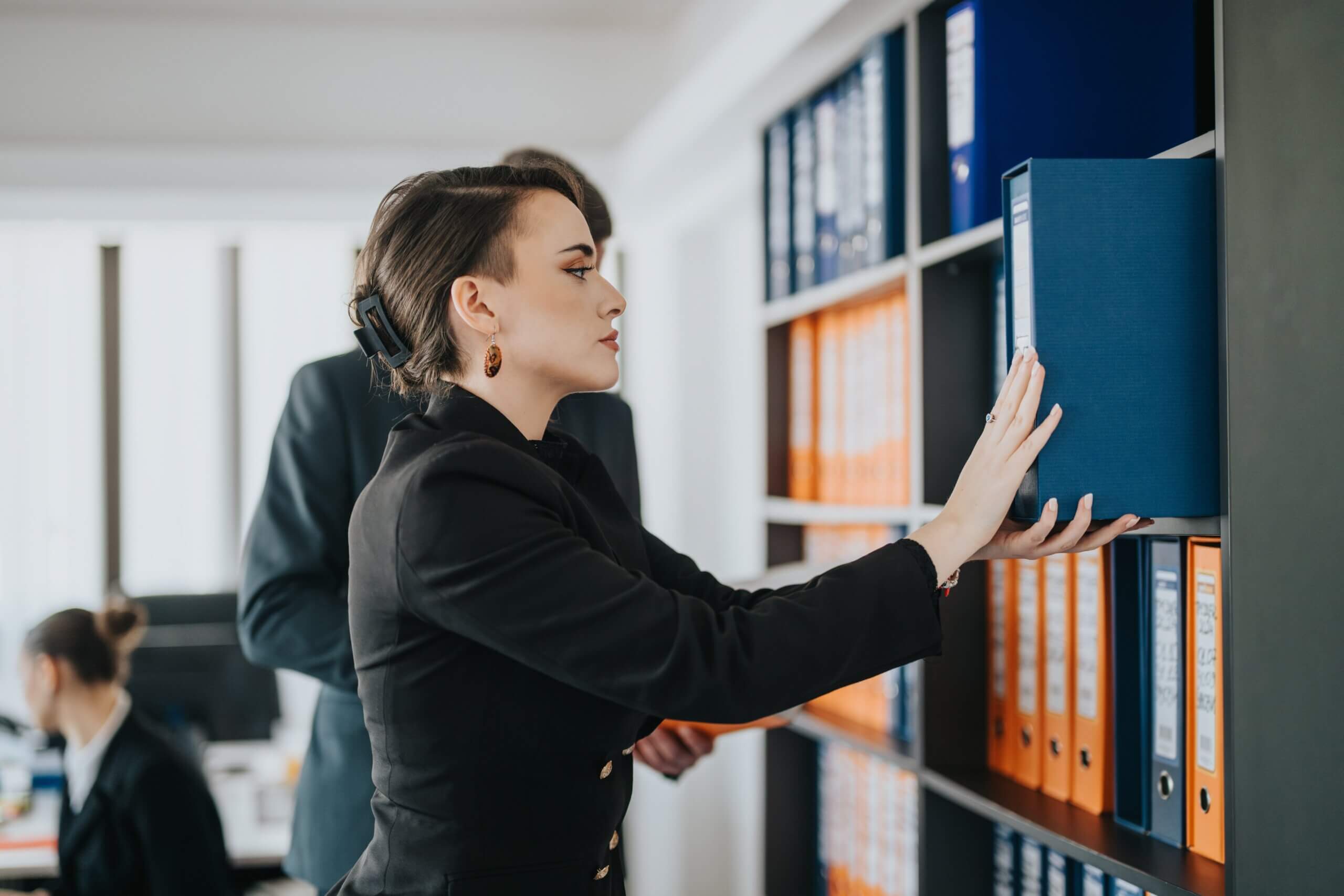 A woman in business attire is reaching for a blue binder on a shelf in an office, with another person standing behind her and more people working at desks in the background.