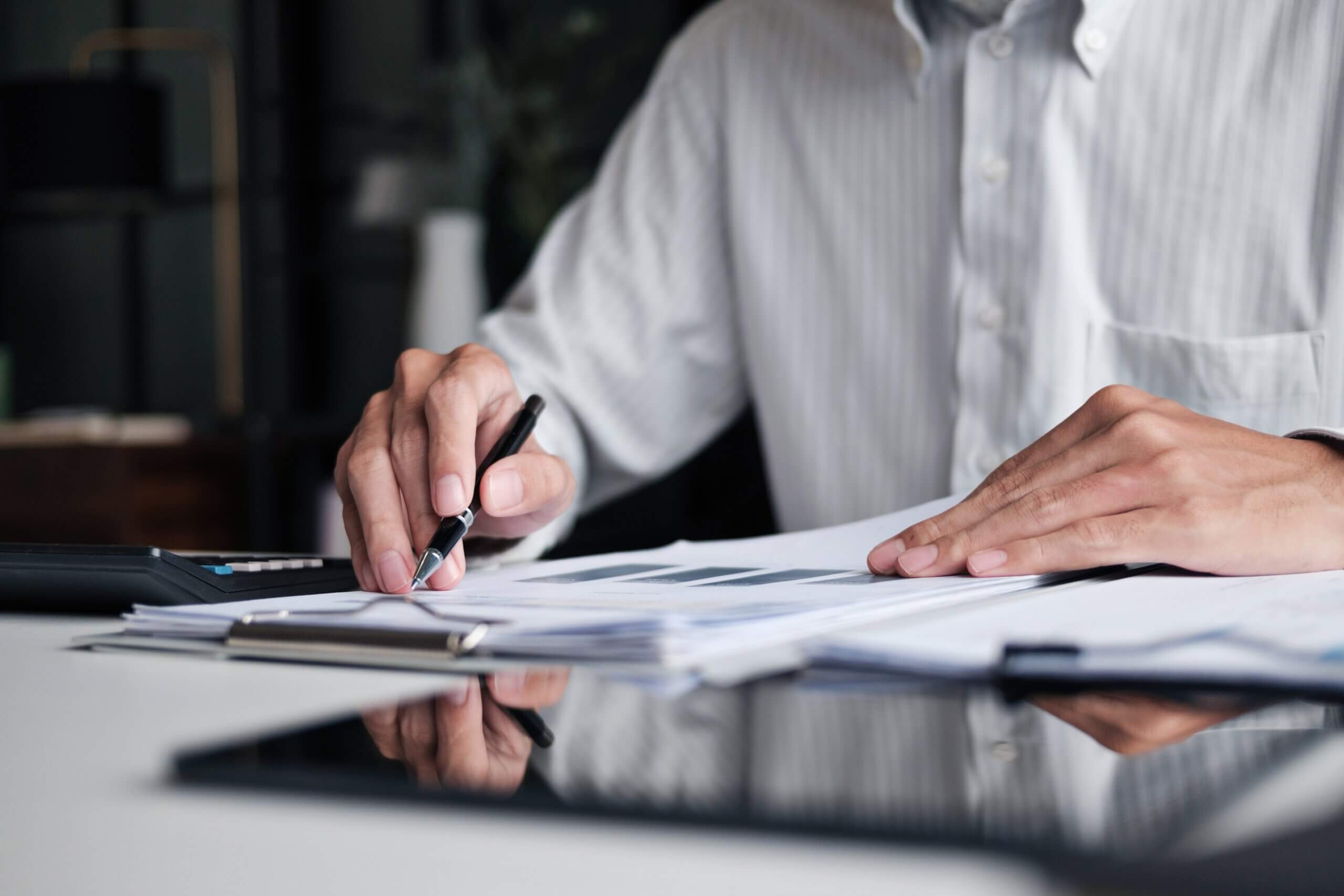 A person in a striped shirt works at a desk, holding a pen and reviewing documents with charts and graphs, with a calculator and a tablet visible in the foreground.