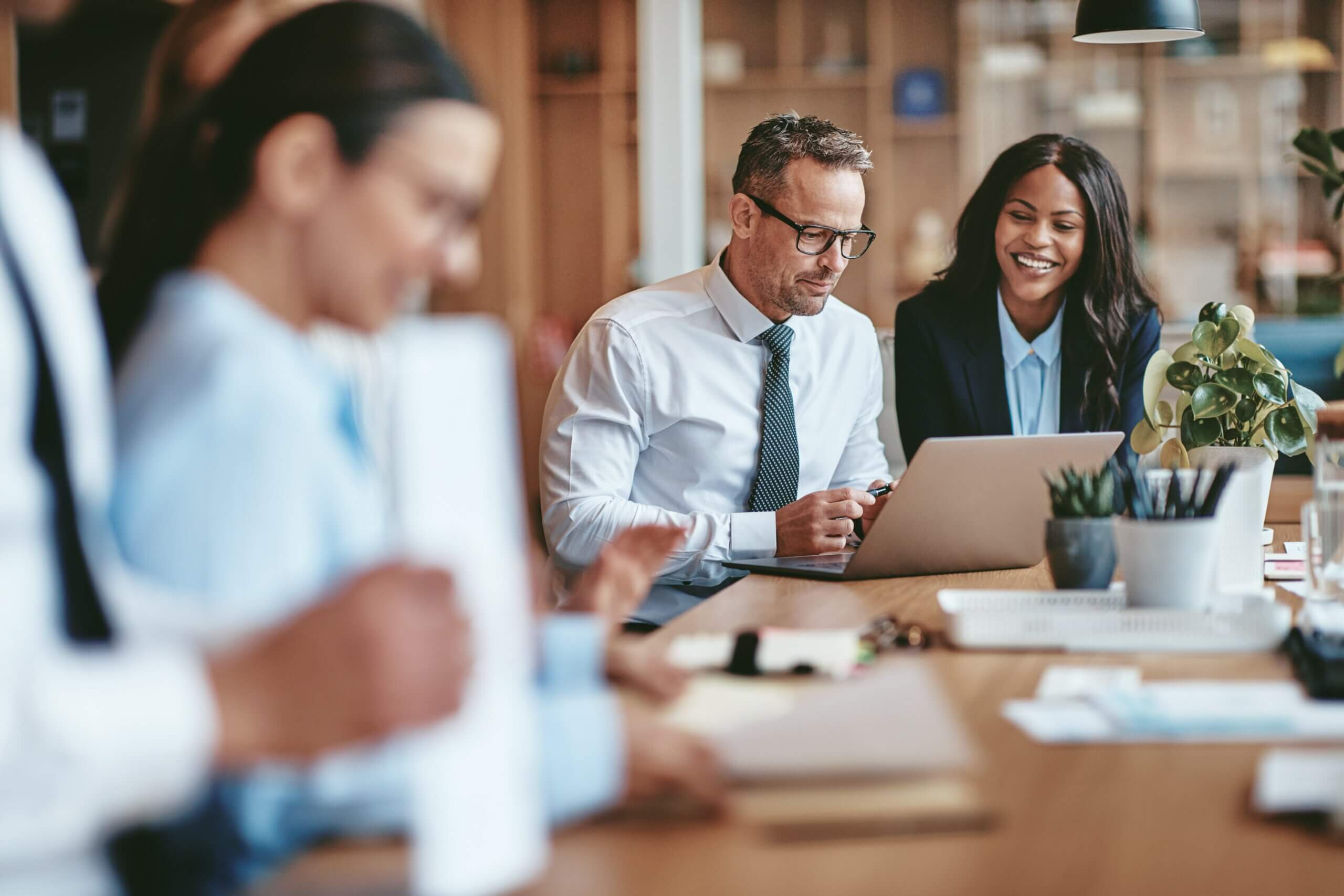 A group of business professionals sits at a table in a modern office, with two colleagues focused on a laptop displaying legal entity identifier data and others working with documents in the foreground.