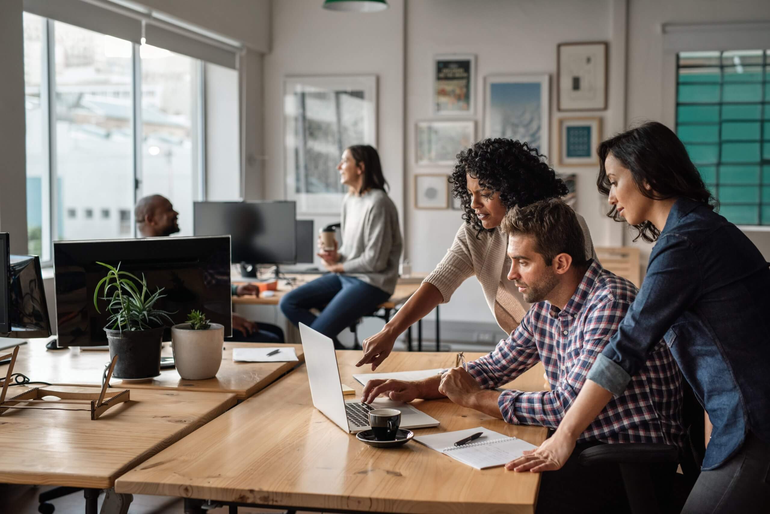 Three coworkers collaborate at a desk, looking at a laptop screen. Two women stand while a man sits, discussing Delaware’s 2025 Business Entity Amendments. In the background, two others chat and drink coffee in a bright, modern office space.