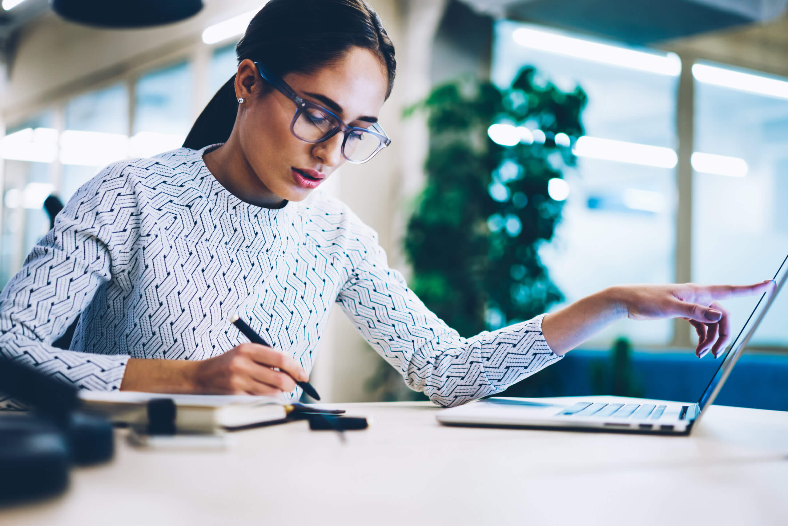 A woman wearing glasses and a patterned white top works at a desk, writing in a notebook with one hand and using a laptop with the other in a modern office setting.