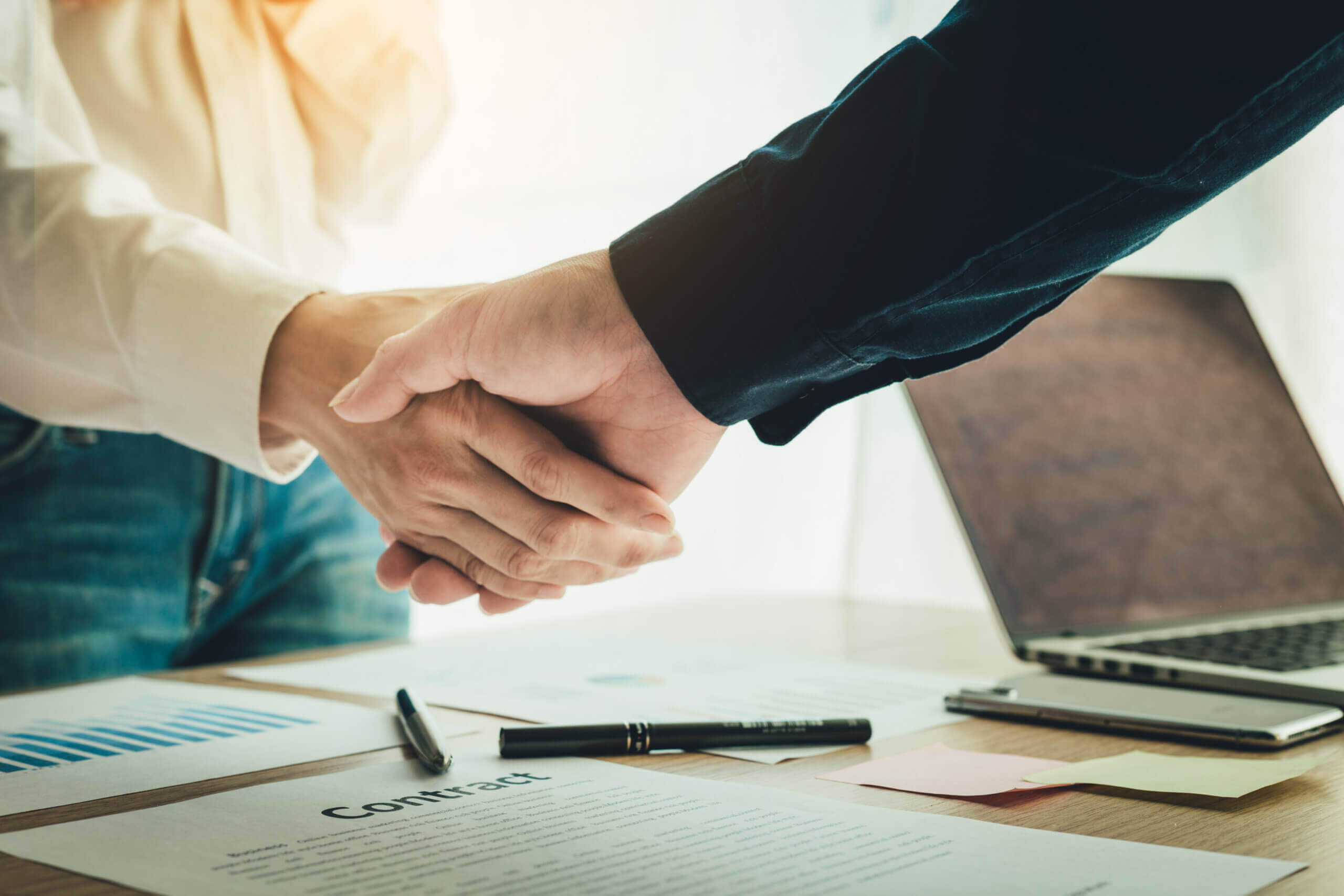 Two people in business attire shaking hands over a desk with documents, a pen, and a laptop, symbolizing a successful agreement or partnership.