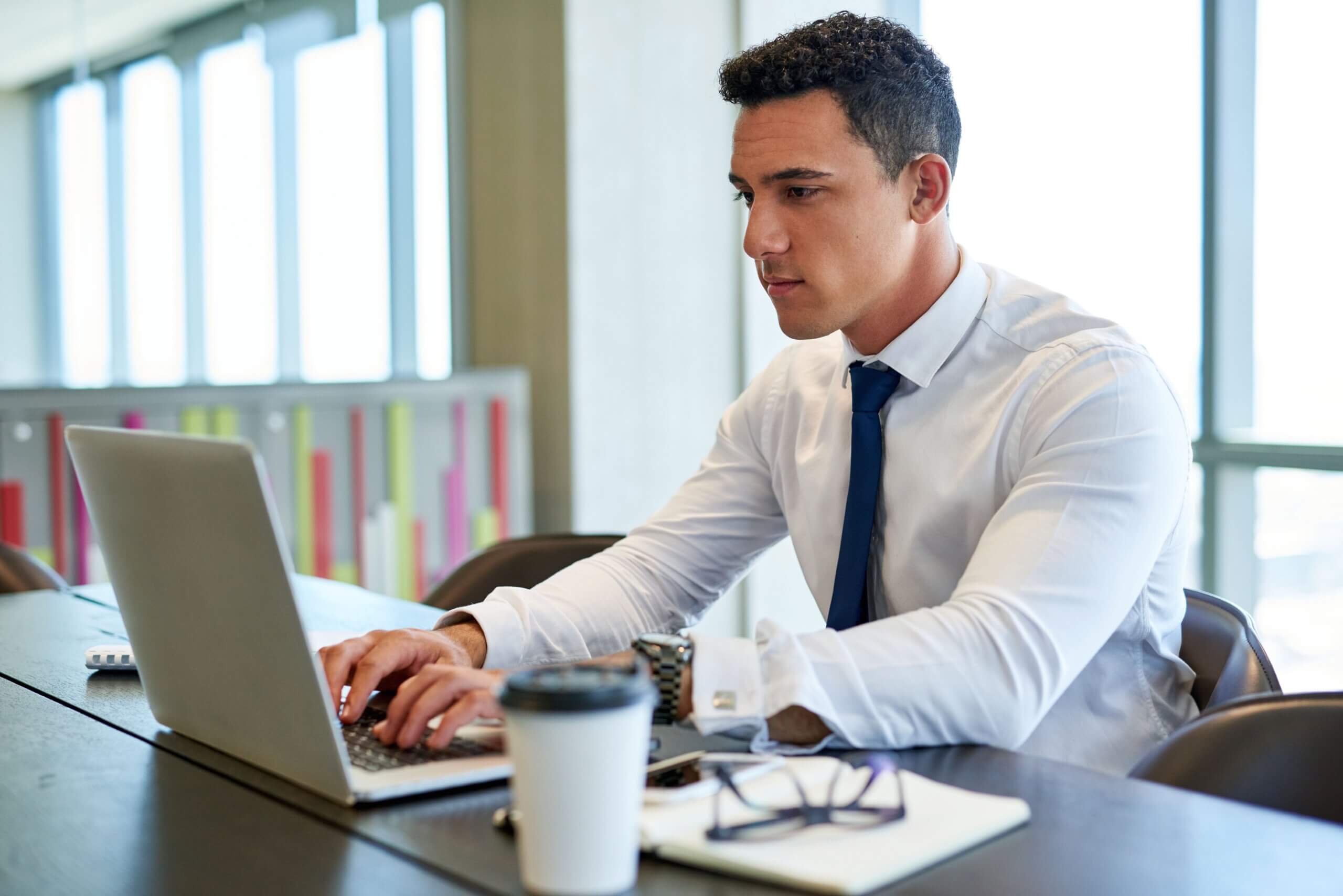 A young man in a white shirt and blue tie works on a laptop at his desk in a modern office, researching Delaware’s 2025 Business Entity Amendments. A coffee cup, notebook, glasses, and pen sit before him as natural light fills the room.