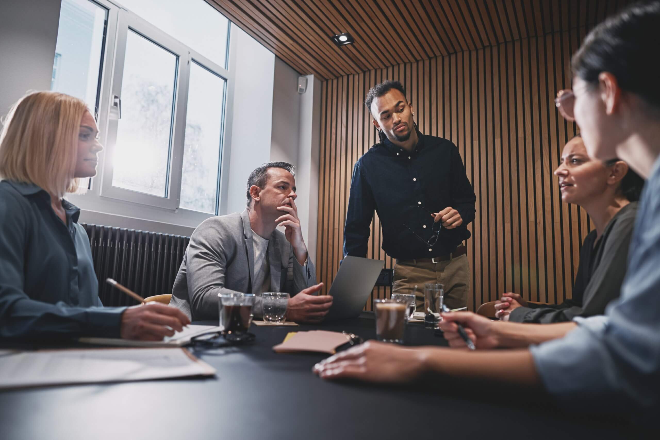 Five people sit and stand around a table in a modern office, engaged in a serious discussion about Delaware’s 2025 Business Entity Amendments. Papers, notebooks, drinks, and a laptop are on the table as sunlight streams through the window.