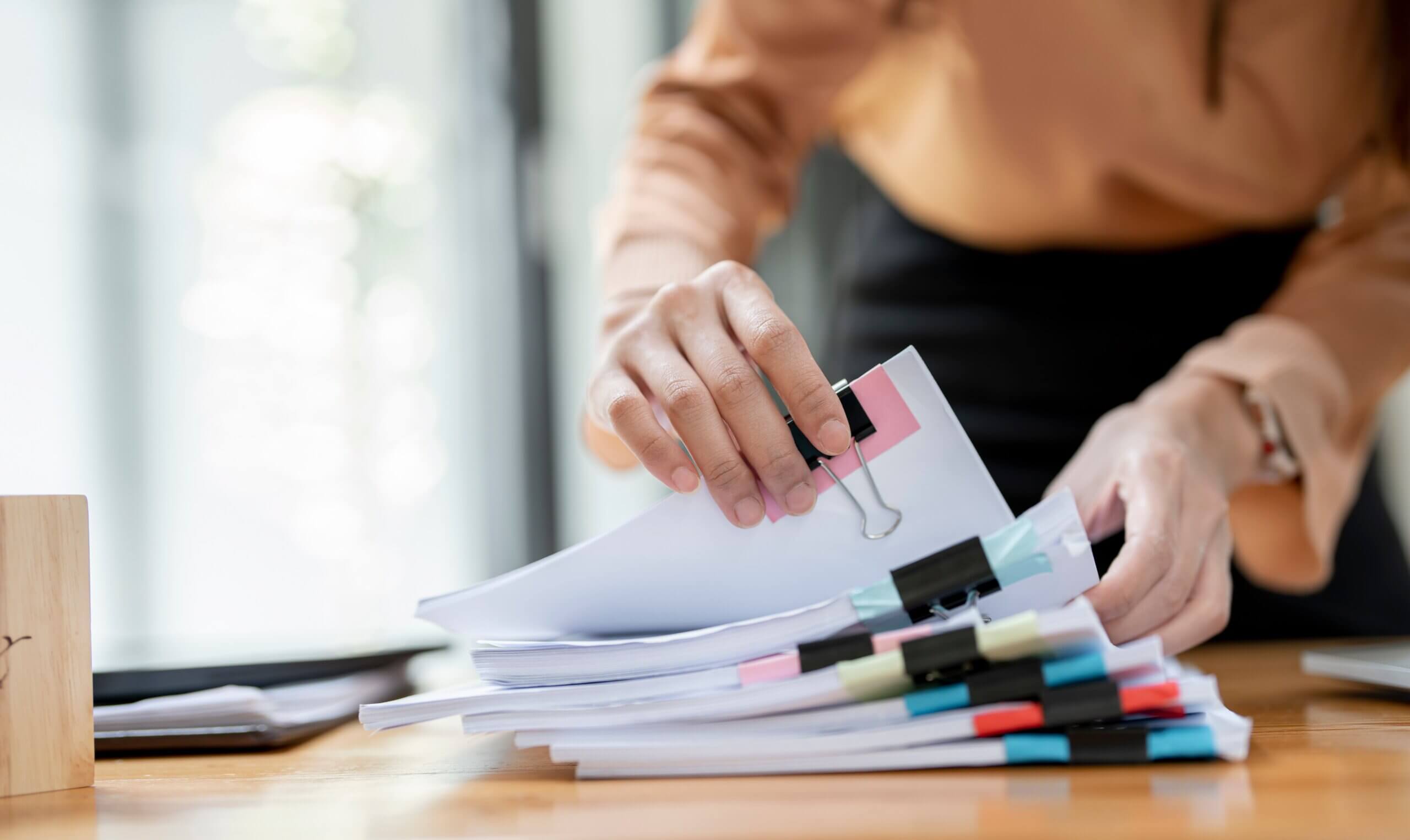 A person organizing a stack of documents with binder clips and colored sticky notes on a wooden desk, with natural light in the background.