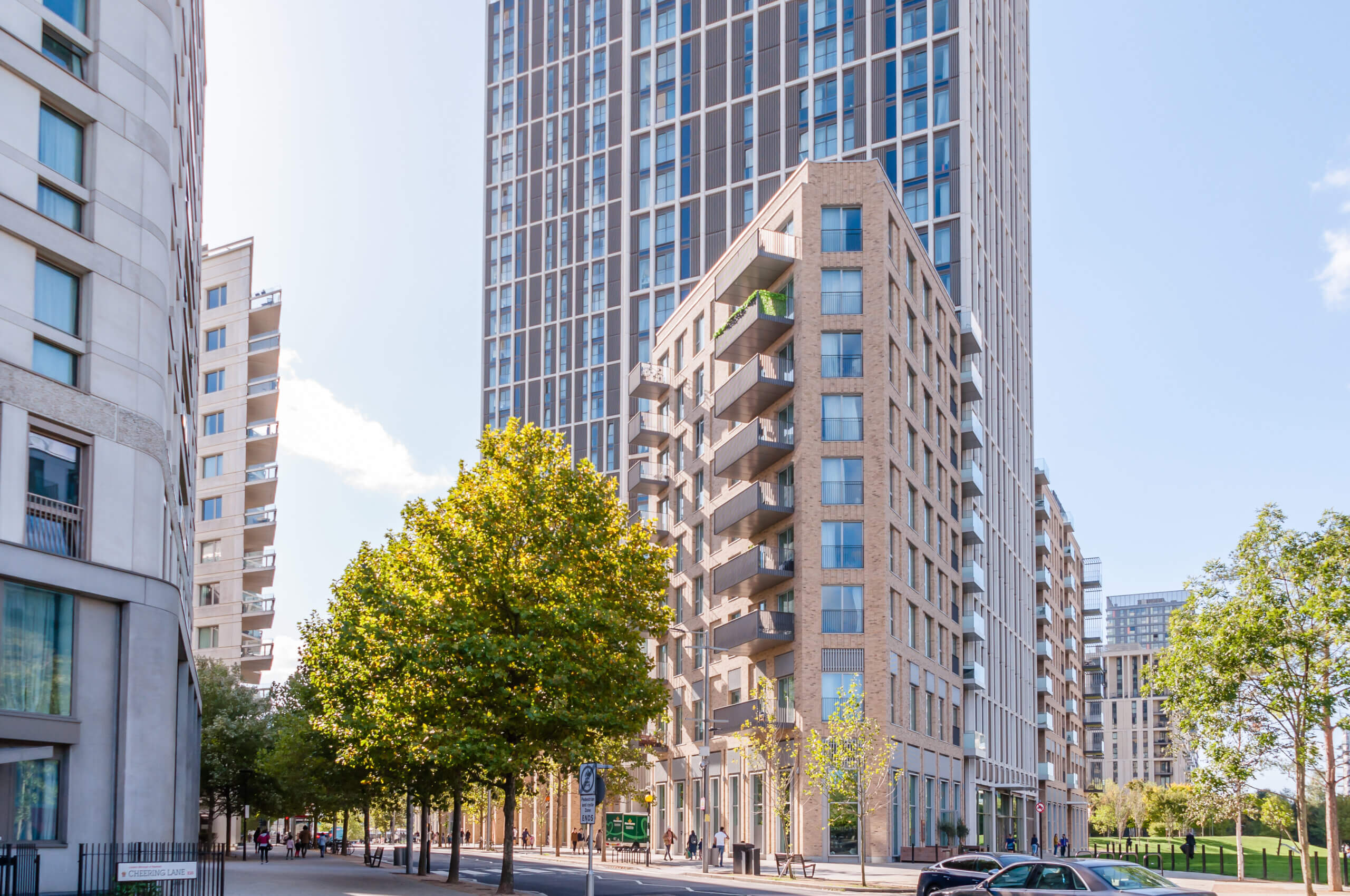 A modern city street lined with trees and tall residential buildings, including a high-rise with a brick facade and balconies. The sky is clear and blue, and there are a few people walking on the sidewalk.
