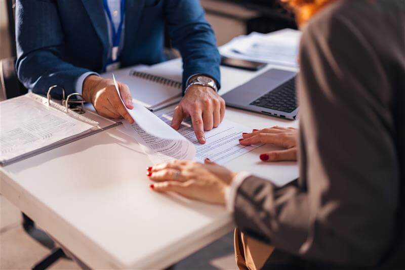 Two people sit at a desk, reviewing documents. One person points to a section on a paper, while the other listens. A laptop and a binder with papers are on the table. Both are wearing business attire.