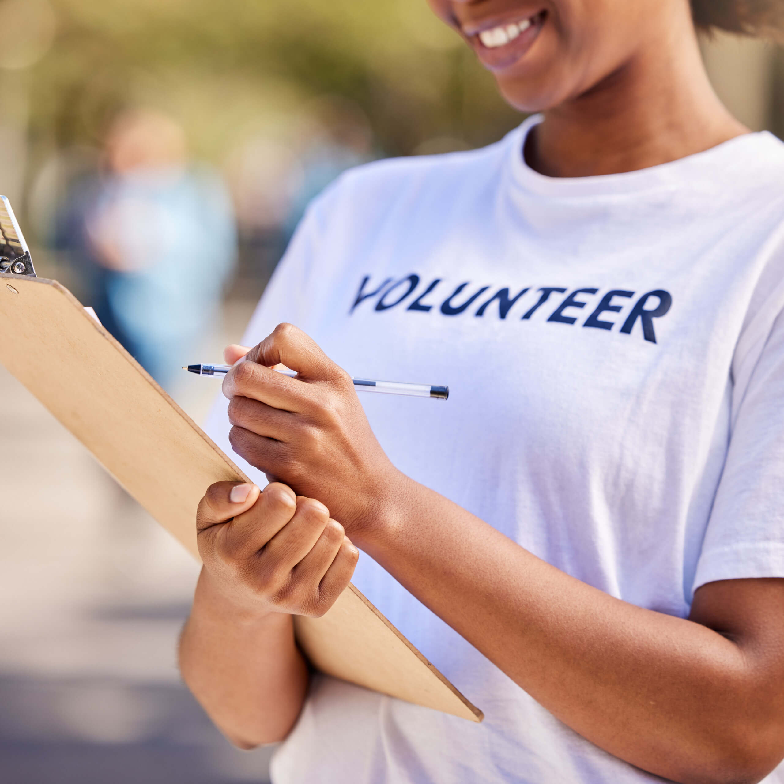 A person wearing a "VOLUNTEER" shirt smiles while holding a clipboard and pen outdoors, ready to take notes or gather information.