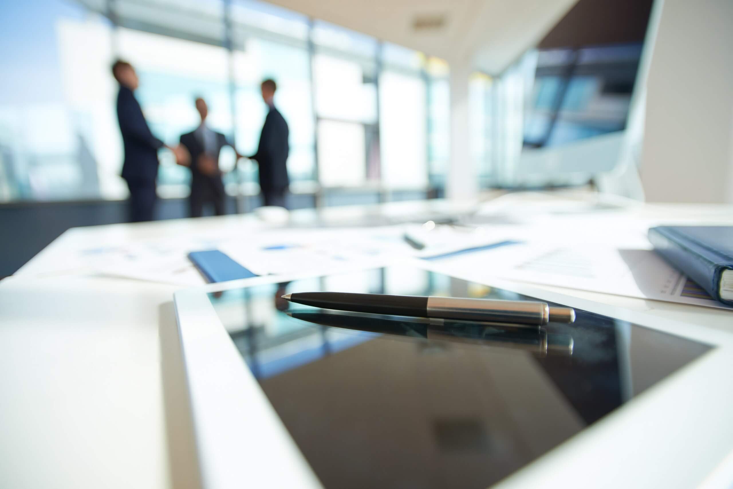 A close-up of a pen resting on a tablet on a desk with documents, while three people in business attire stand and talk in the blurred background of a bright office.