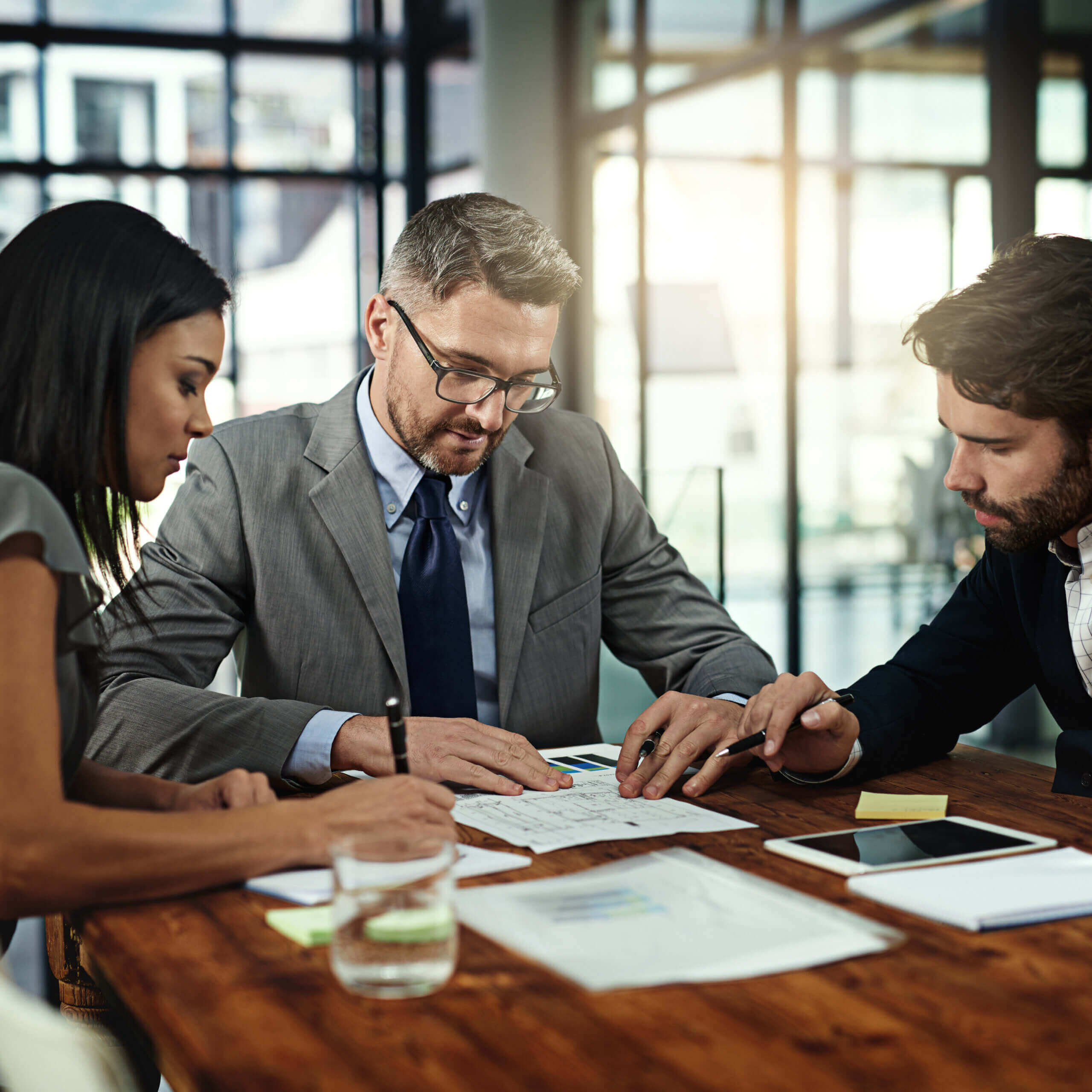 Three business professionals sit around a wooden table reviewing documents together in a bright, modern office, with a tablet, papers, and a glass of water in front of them.