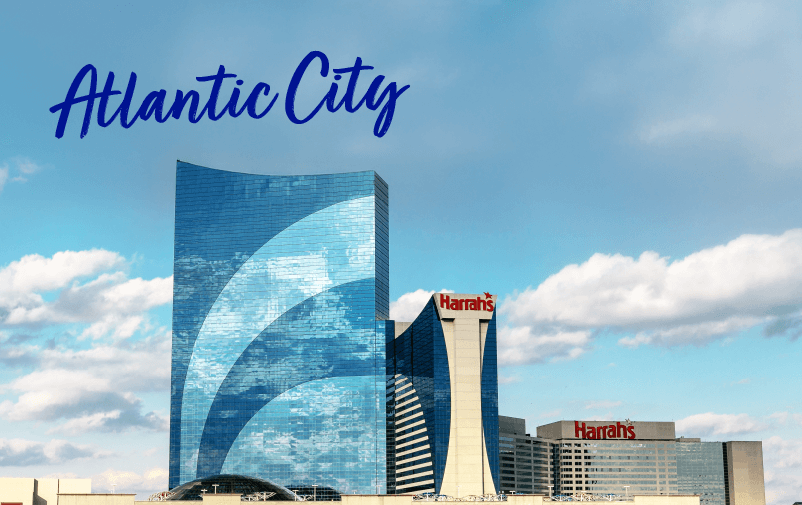 Tall modern buildings and casinos in Atlantic City, with “Atlantic City” written in blue script across the sky. Harrah’s casino signs are visible on multiple buildings—a perfect backdrop for upcoming lending conferences 2025 under a partly cloudy sky.