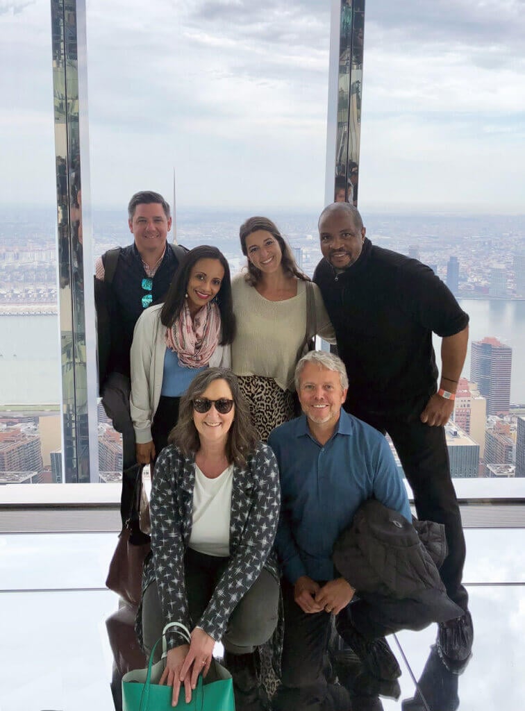 Six people pose together inside a glass observation deck high above a city skyline. Smiling at the camera, they reflect the teamwork and professionalism that define Cogency Global’s corporate compliance solutions.