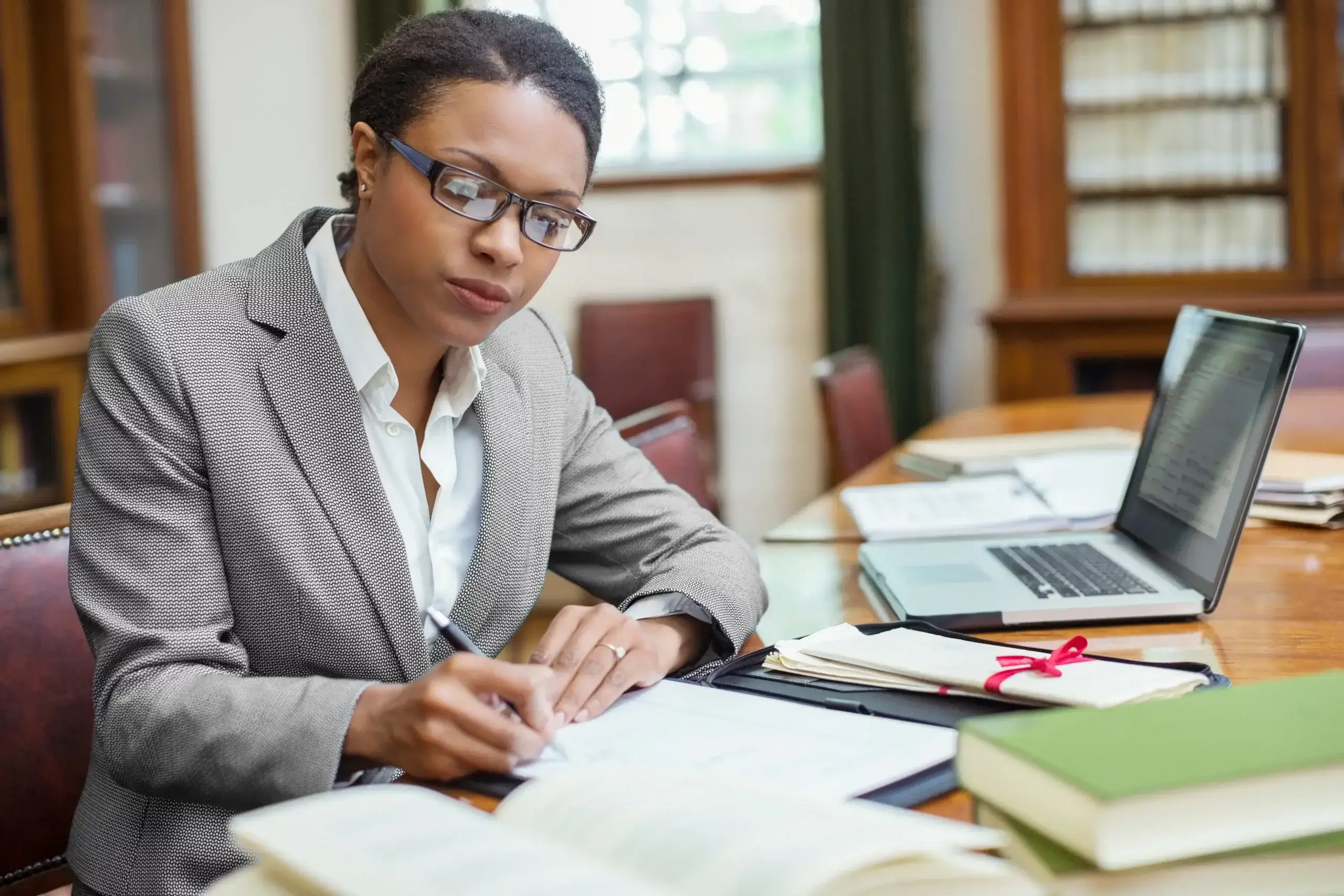 A woman in a gray suit sits at a desk in a library or office, writing in a notebook while conducting international due diligence. She wears glasses, with a laptop, open books, and documents spread out before her.