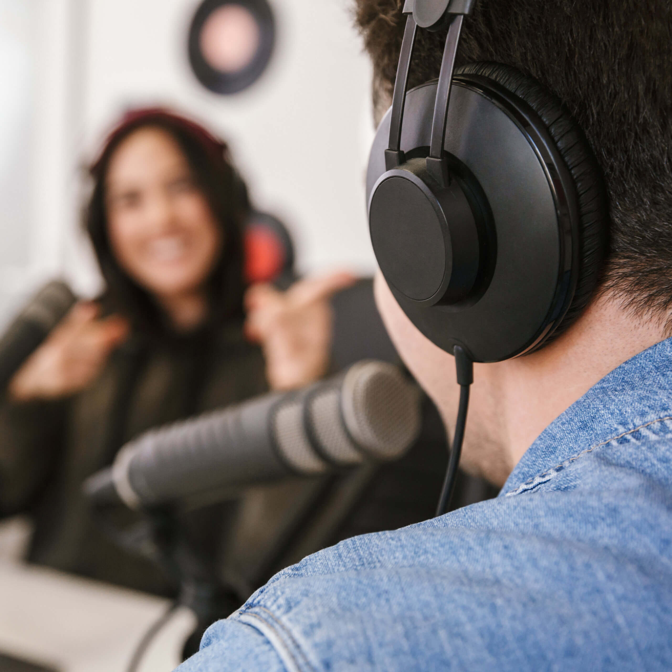 A person wearing headphones and a denim jacket speaks into a microphone in a recording studio, while another person in the background gestures and smiles, both appearing engaged in conversation.