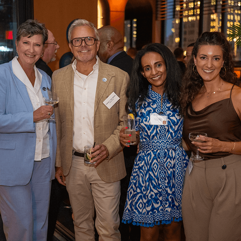 Four people dressed in semi-formal attire smile and pose together at an indoor social event, each holding a drink. City lights are visible through the windows in the background.
