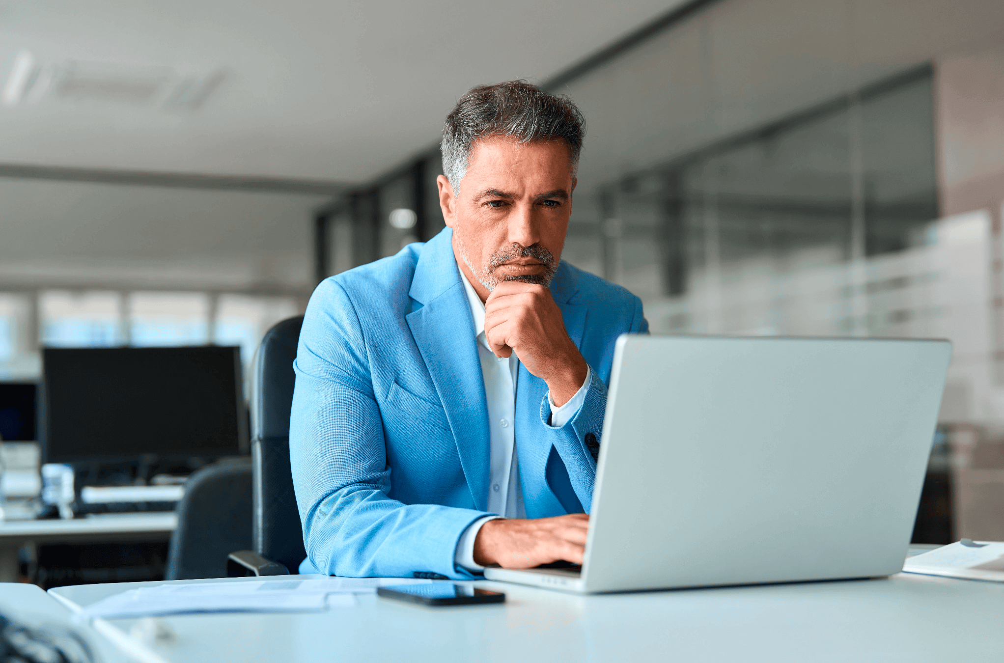 A man in a light blue blazer sits at a desk in an office, looking thoughtfully at his laptop screen with his hand resting on his chin, as he reviews the identity verification process.