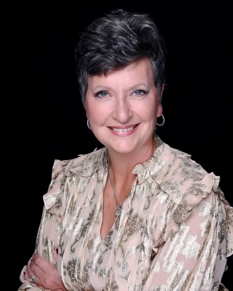 A woman with short, dark hair and blue eyes smiles while wearing a patterned, light-colored blouse and hoop earrings, against a black background.