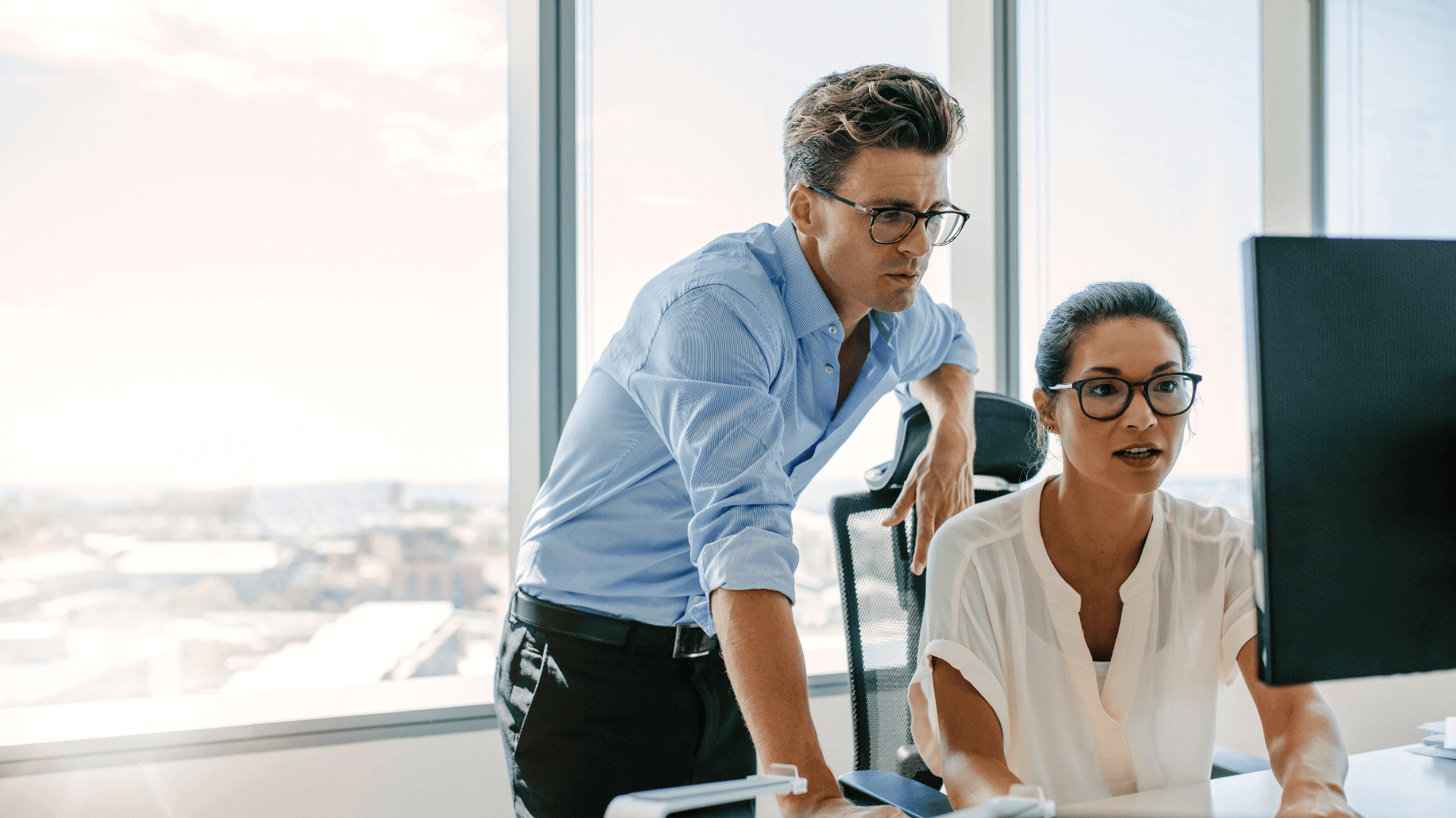 A man and a woman in business attire work together at a computer in a bright office with large windows overlooking a cityscape. The man stands beside the woman, both focused on the monitor.