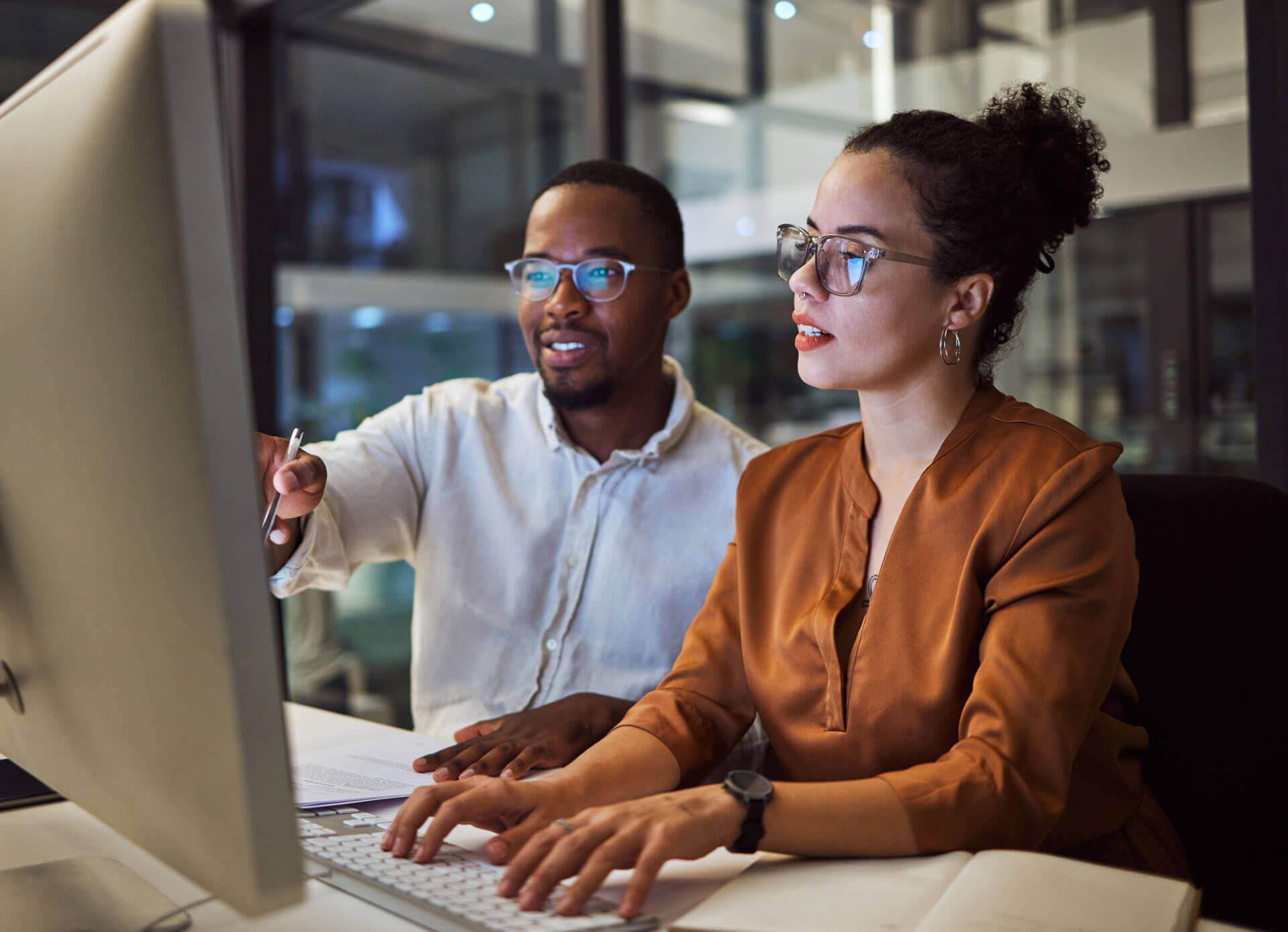 A man and a woman work together at a computer in an office setting. The man points at the screen while the woman types on the keyboard, illustrating Cogeny Global’s name reservation services. Both are wearing glasses and are focused on the monitor.