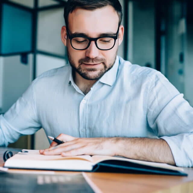 A man wearing glasses and a light blue shirt sits at a desk, writing in a notebook with a pen. He appears focused and is surrounded by office materials in a modern workspace.