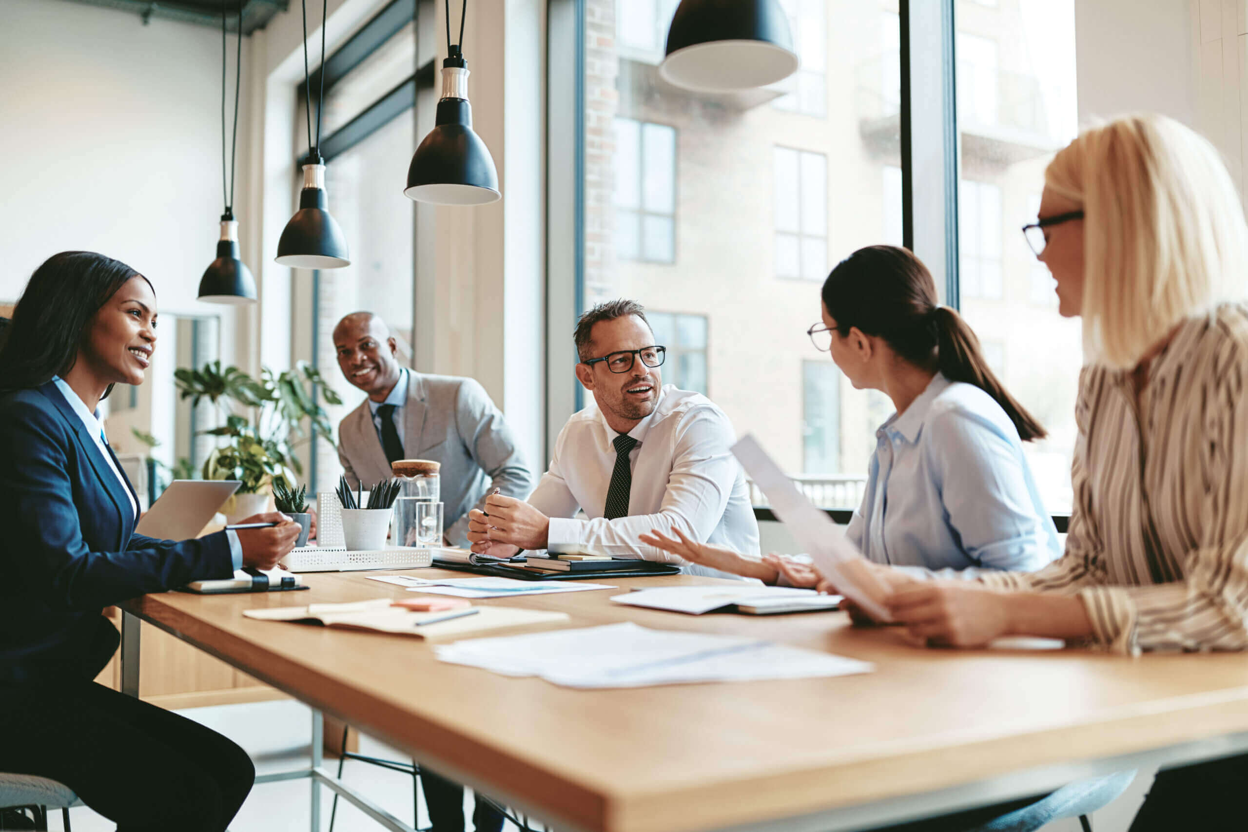 A group of five people in business attire is engaged in a meeting around a wooden table in a modern office, discussing Cogency’s Global’s corporate services. Papers and devices are on the table, and large windows reveal an urban background.