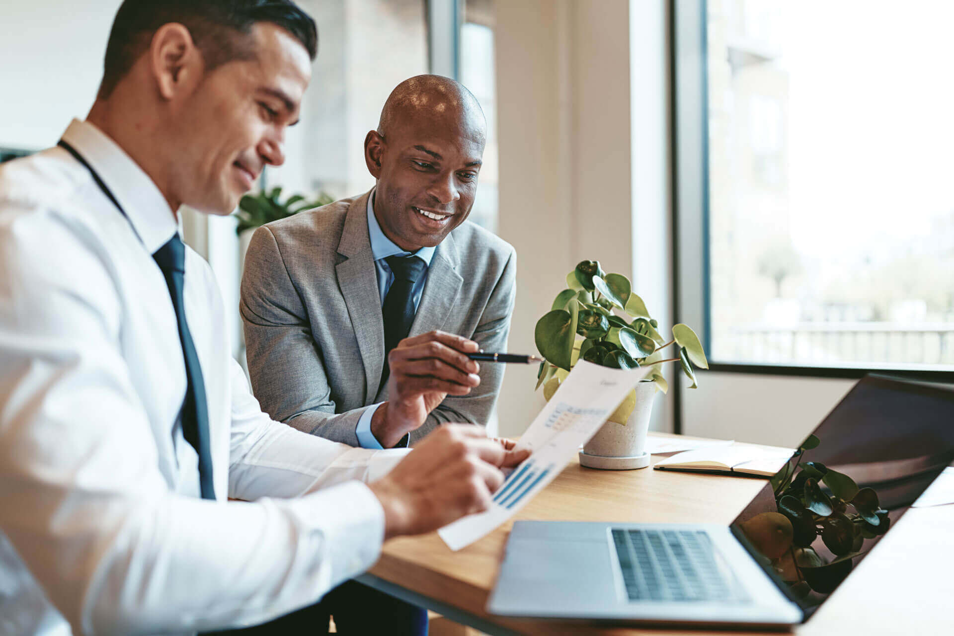 Two businessmen in a meeting room, one holding a document with graphs. They are engaged in discussion, seated at a wooden table with a laptop and a potted plant, representing Cogeny Global’s nonprofit sales tax exemptions services.