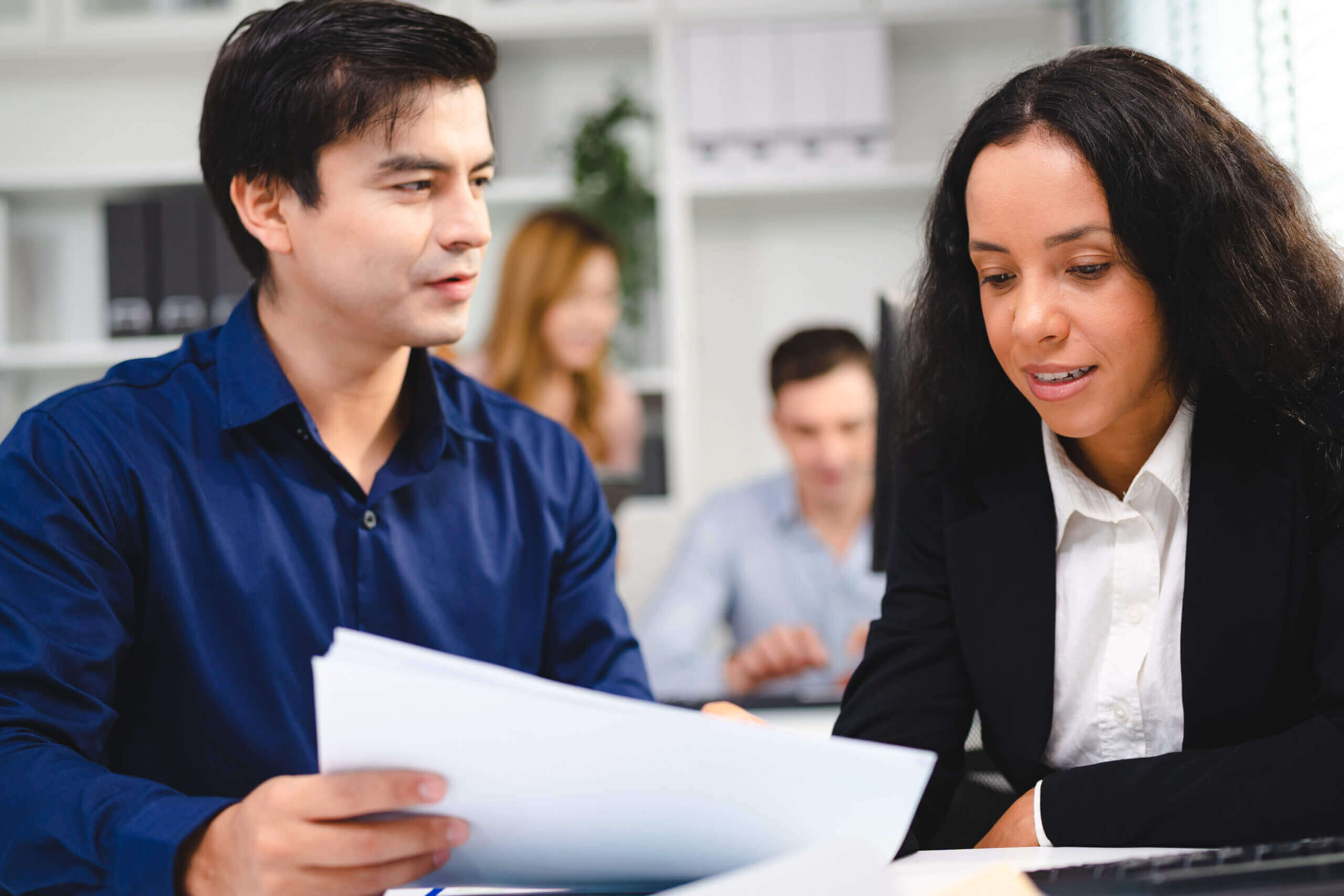 A man and a woman in business attire review documents together at a desk in an office, with two colleagues working in the background.
